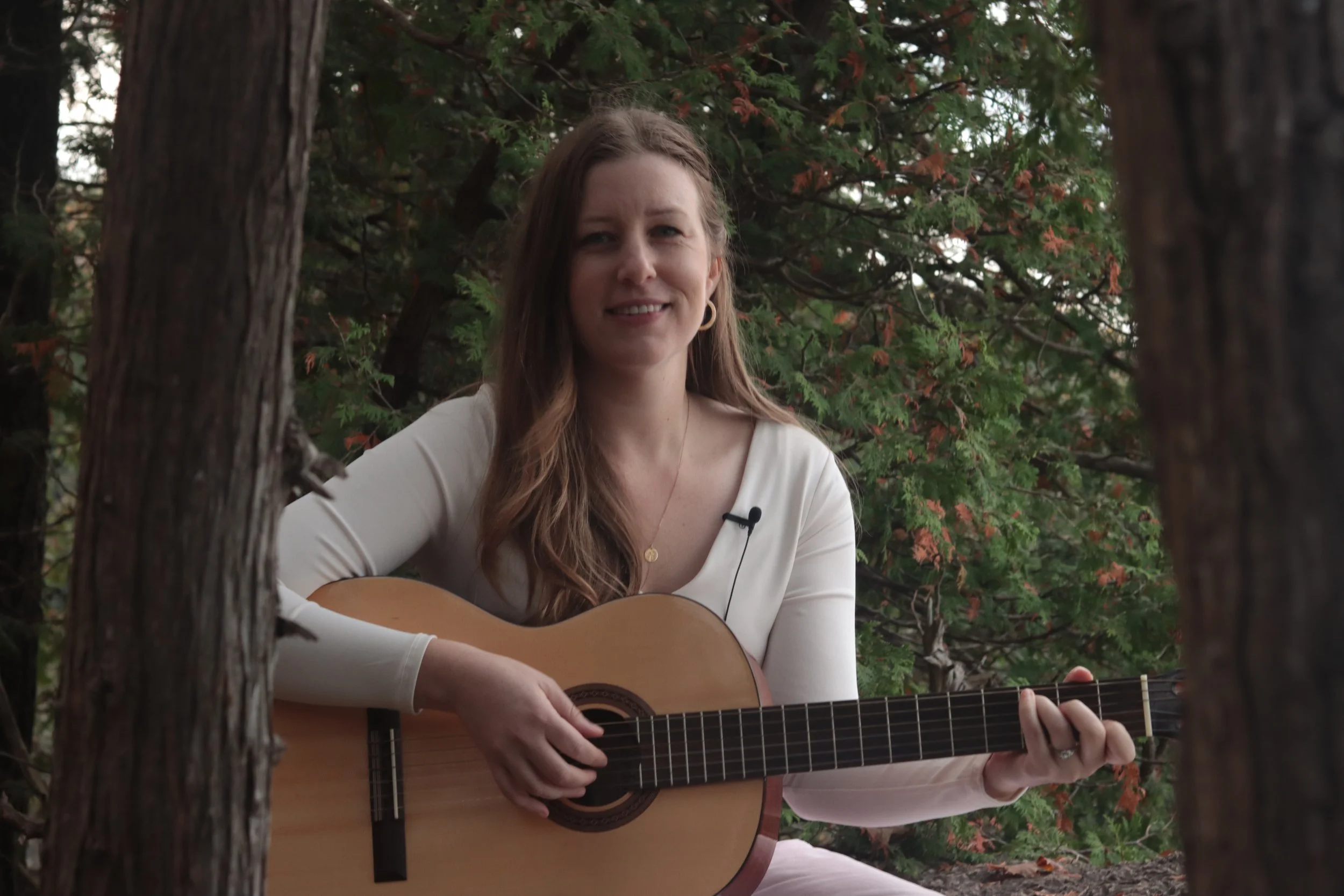 A woman with long brown hair wearing a white top, sitting outdoors among trees, playing an acoustic guitar and smiling at the camera.