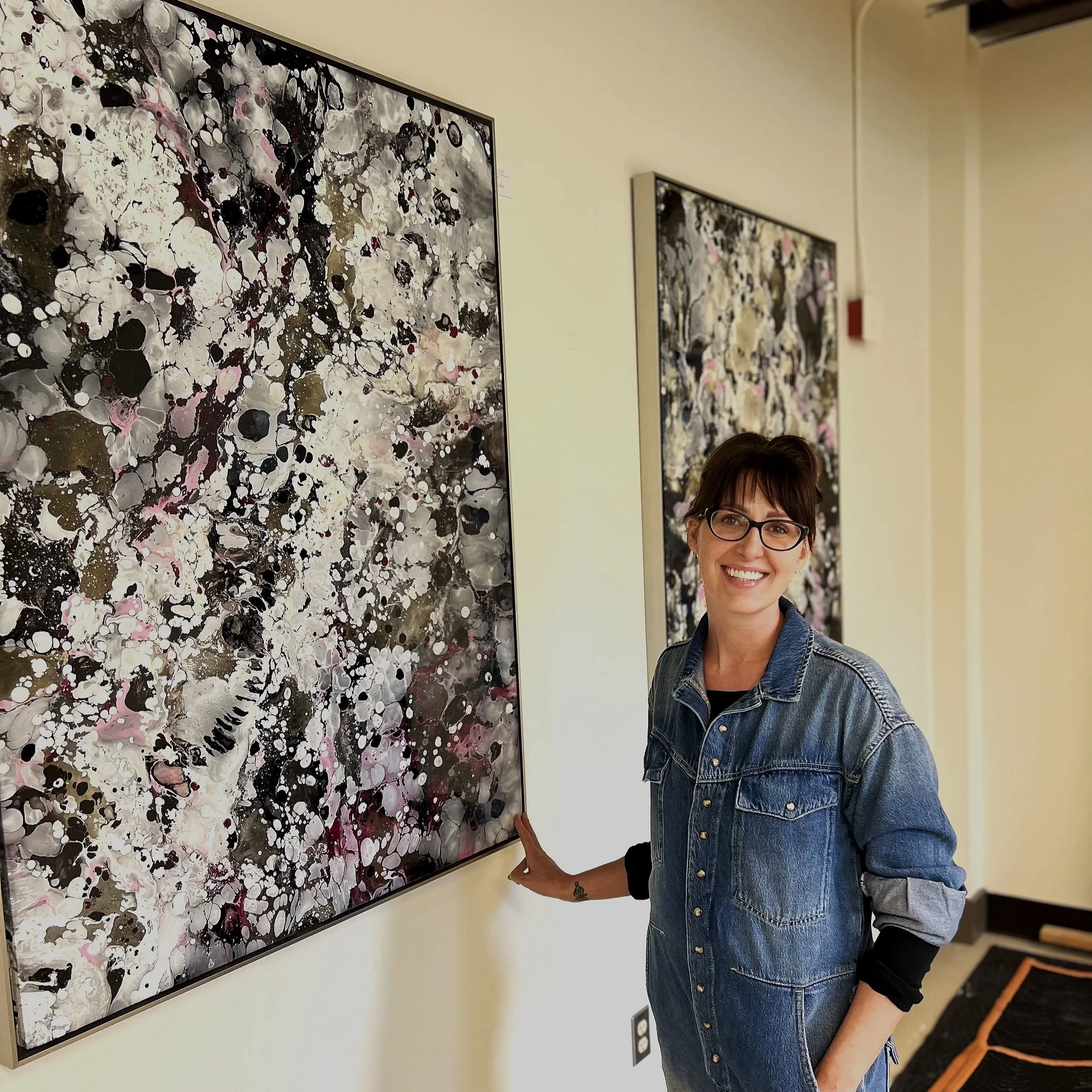 A woman with short hair and glasses smiling and standing in front of abstract artworks hanging on a gallery wall. She is wearing a denim jacket and jeans.