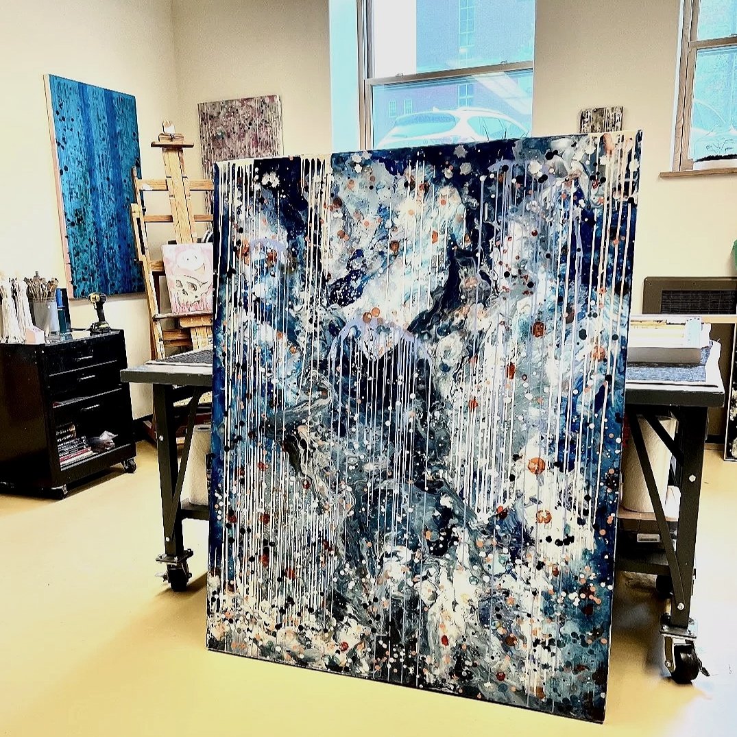 An abstract painting leaning against a table on a yellow floor in an art studio, with other artwork and art supplies visible in the background.