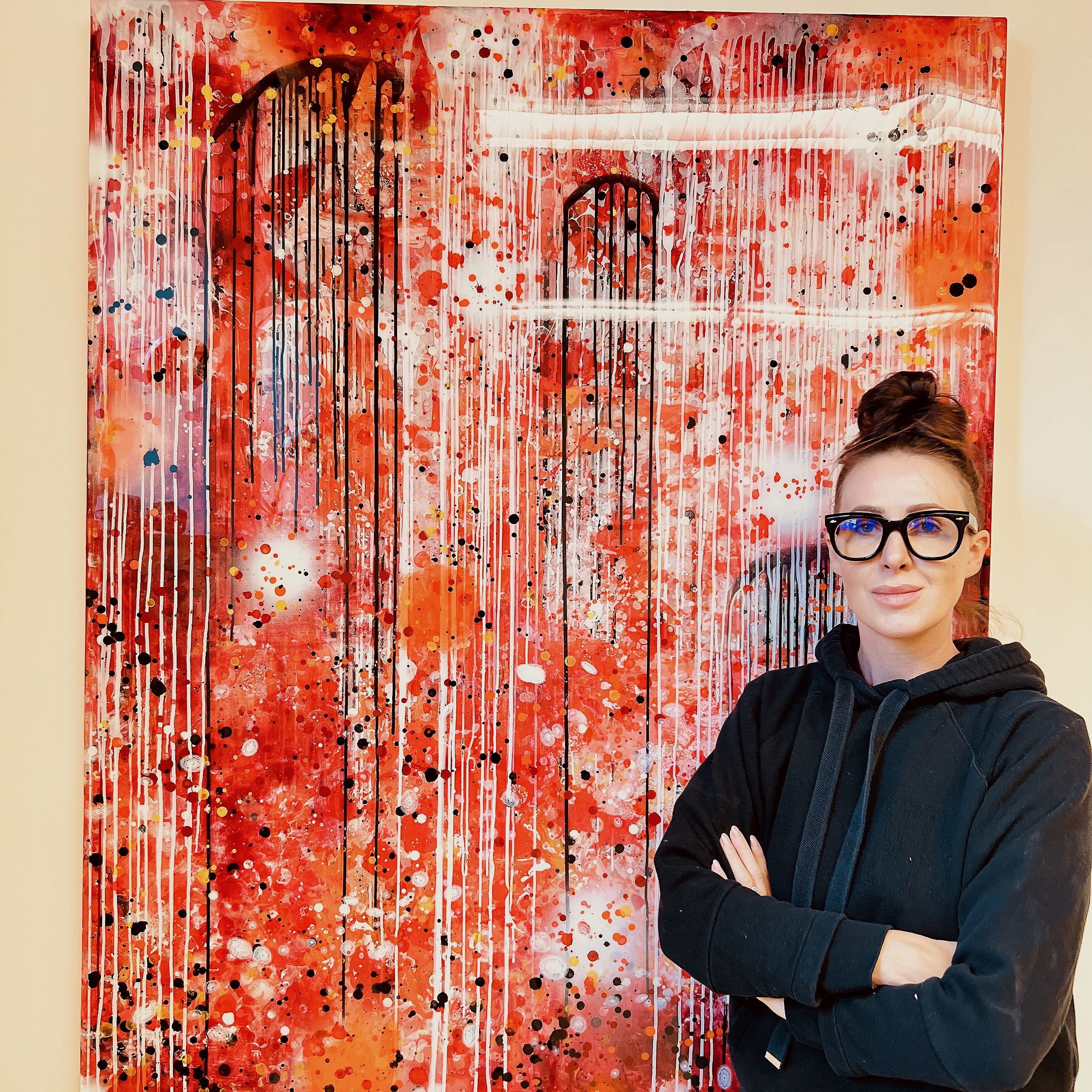 A woman wearing glasses and a black hoodie standing arms crossed in front of an abstract red, white, and black splatter painting.