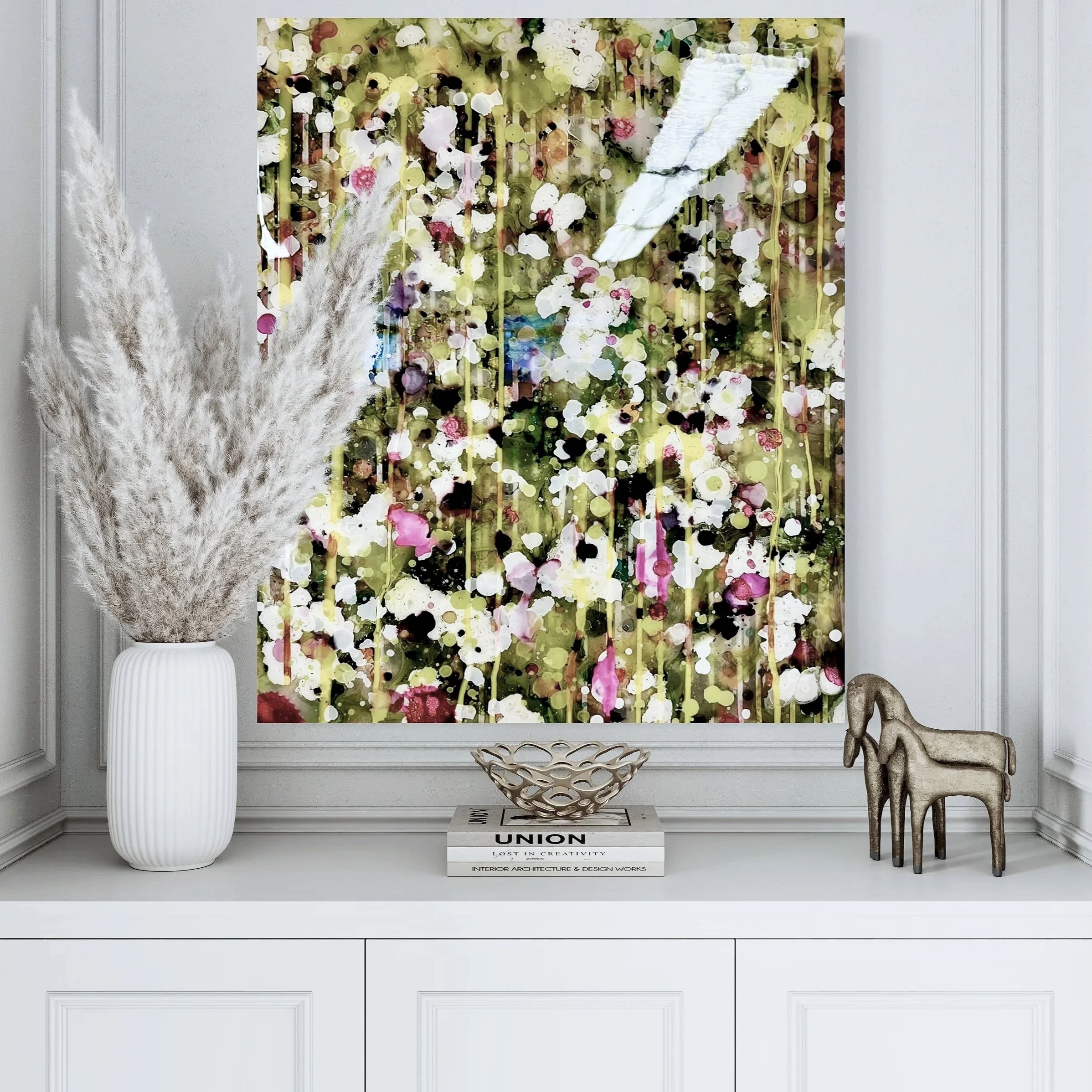 Decorative wall art with colorful abstract design, placed above a white dresser. To the left, a tall white vase with pampas grass. On the right, small animal-shaped stools and a stack of books with a gold decorative bowl on top.