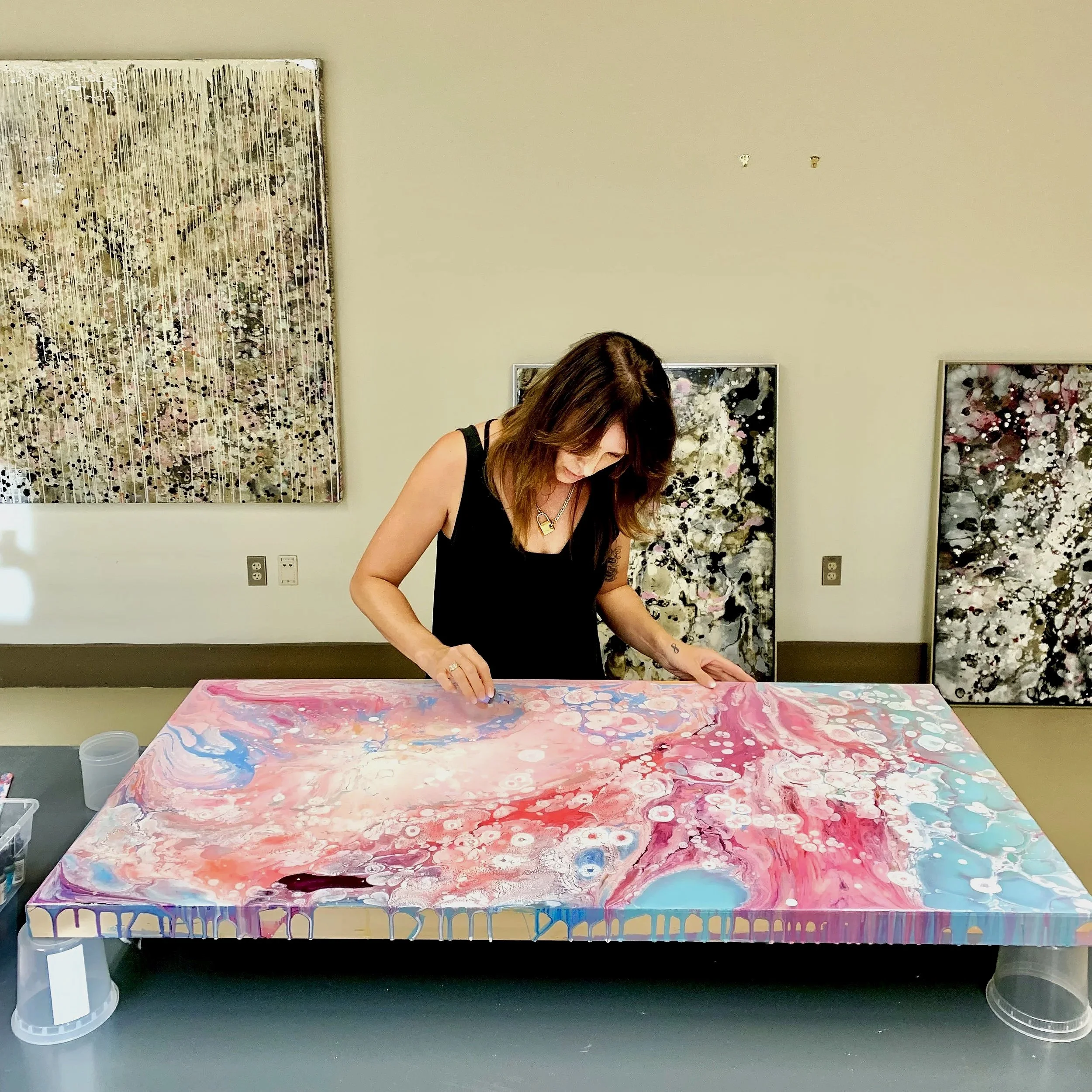 A woman with brown hair and a black sleeveless top is working on a colorful abstract painting on a table. The background has beige walls with three framed artworks and a large piece of abstract art, featuring black, white, pink, and other colors. The