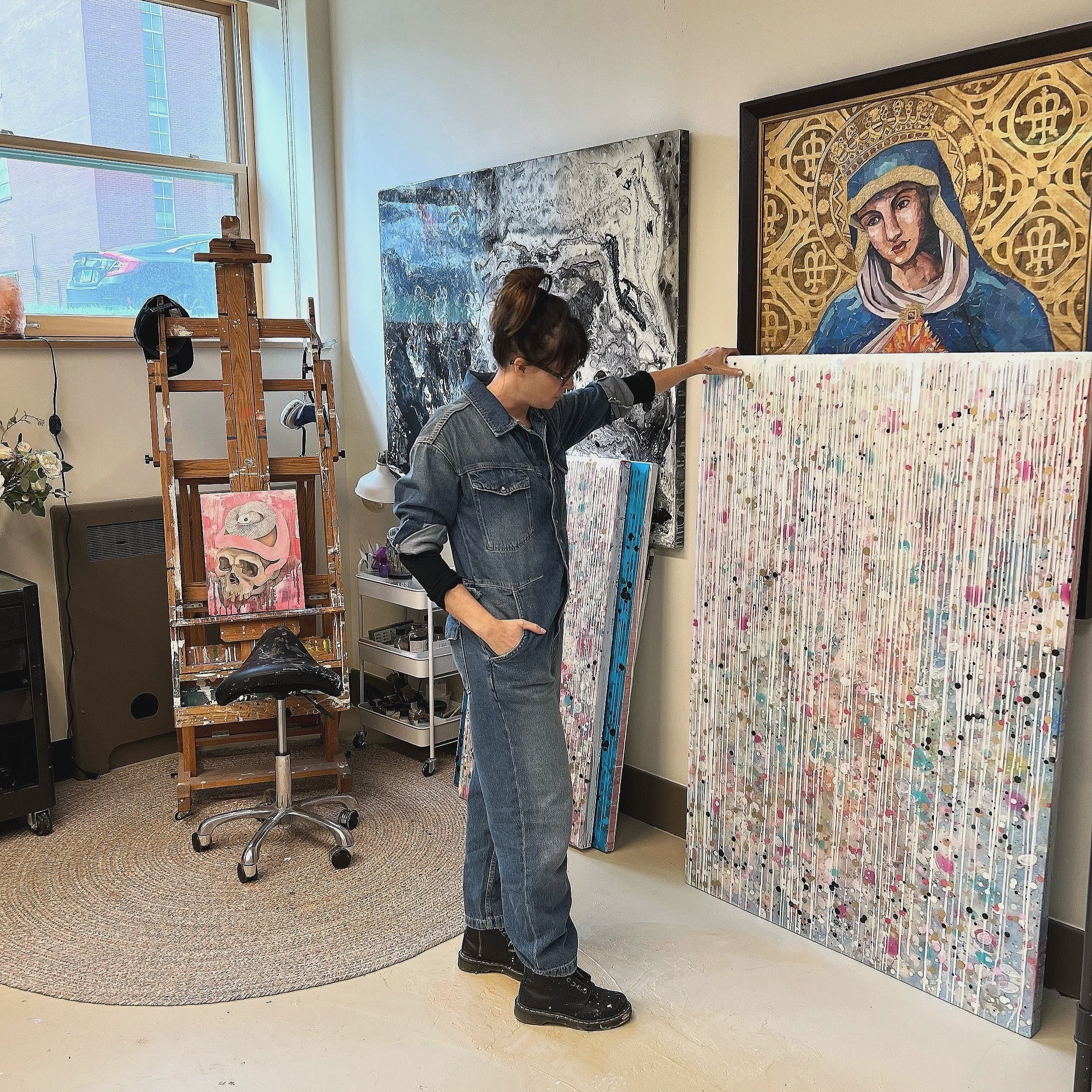 An artist in a denim jacket and black boots looks at a large colorful abstract painting in an art studio. The studio has a window with a car parked outside, a wooden easel with a skull painting, and several other artworks on the wall.