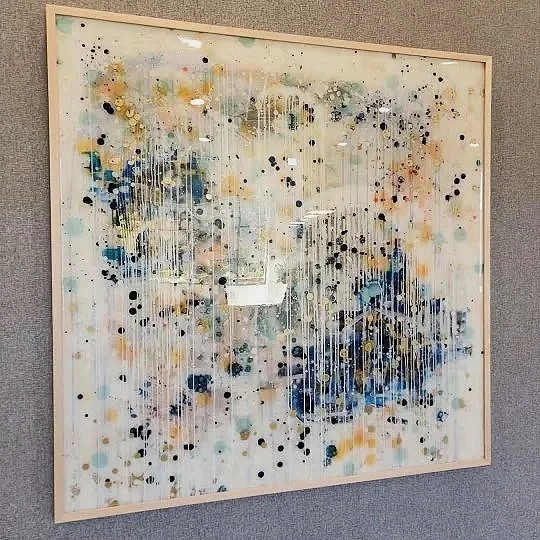 Abstract colorful painting with drips and splatters of paint in blue, black, yellow, and orange on a white background, framed and hanging on a gray wall.