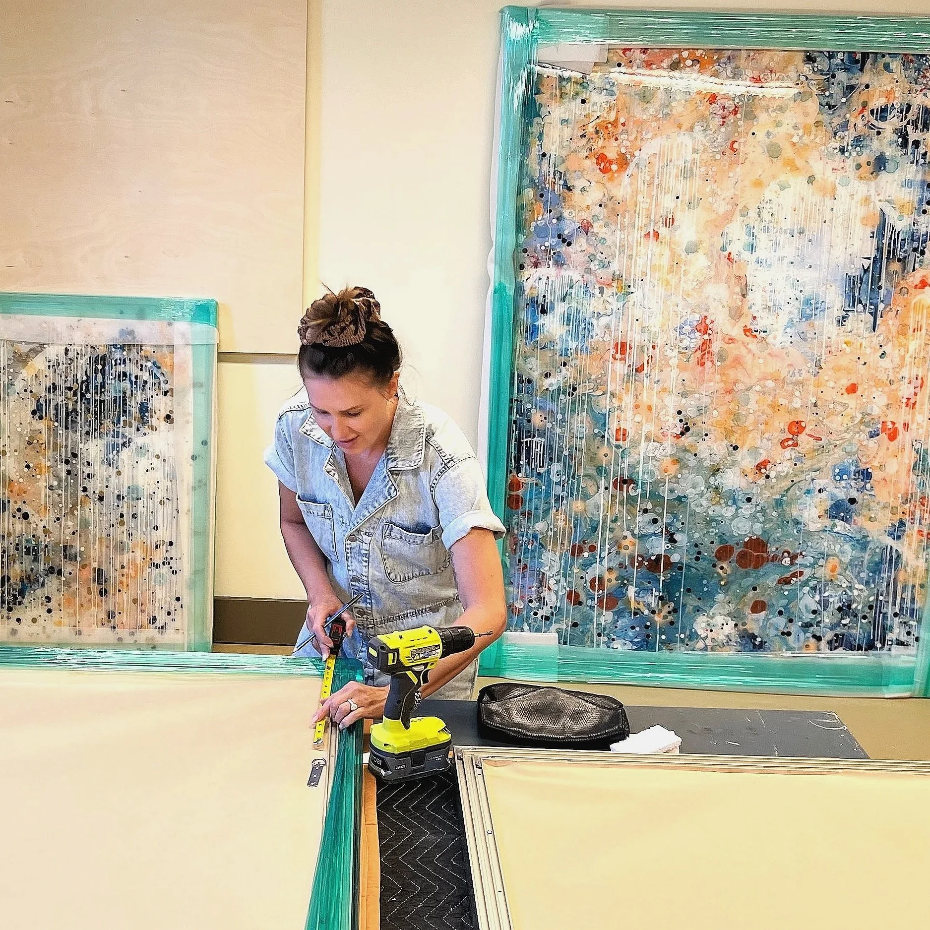 A woman with her hair tied up working on a project with a power drill and ruler in a room with colorful abstract artwork on the walls.
