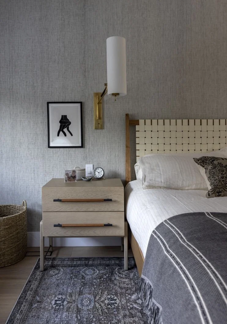 A modern side table sits besides a modern bed with a woven headboard in a moody bedroom.