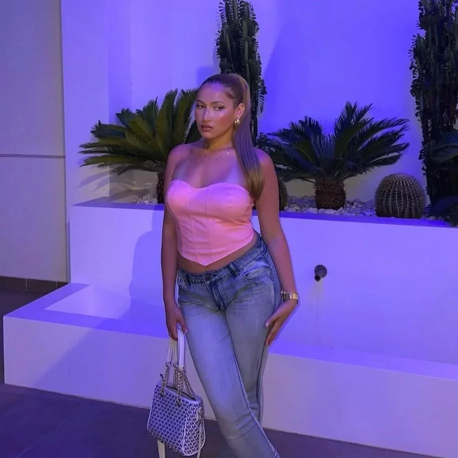 A woman standing in front of a decorative indoor garden with palm and cactus plants, illuminated by purple lighting. She is wearing a pink strapless top, jeans, and holding a patterned handbag.