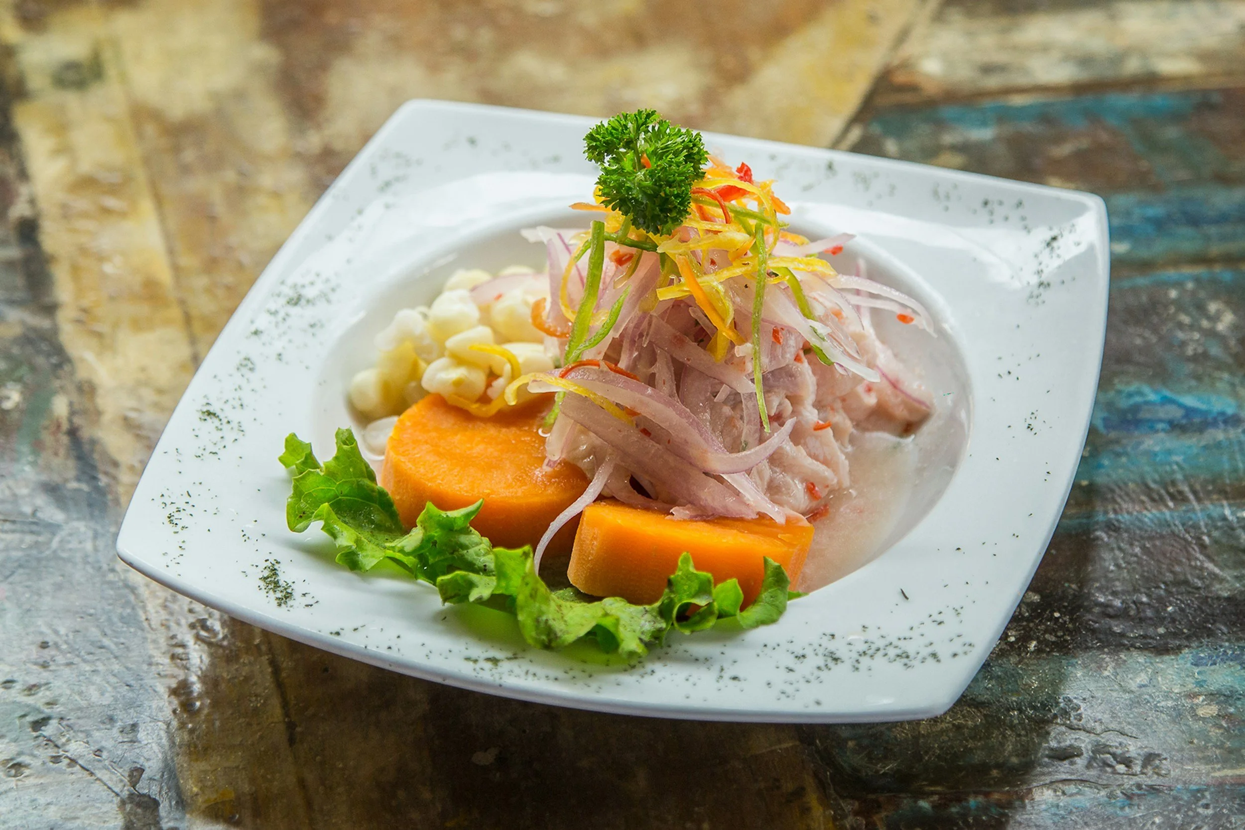 A plate of ceviche with sliced sweet potatoes, corn, and garnished with cilantro and shredded vegetables on a decorative white square plate.
