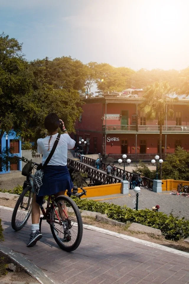 Person in school uniform standing with a bicycle, taking a photo of a building across a small street in a town square during sunset.