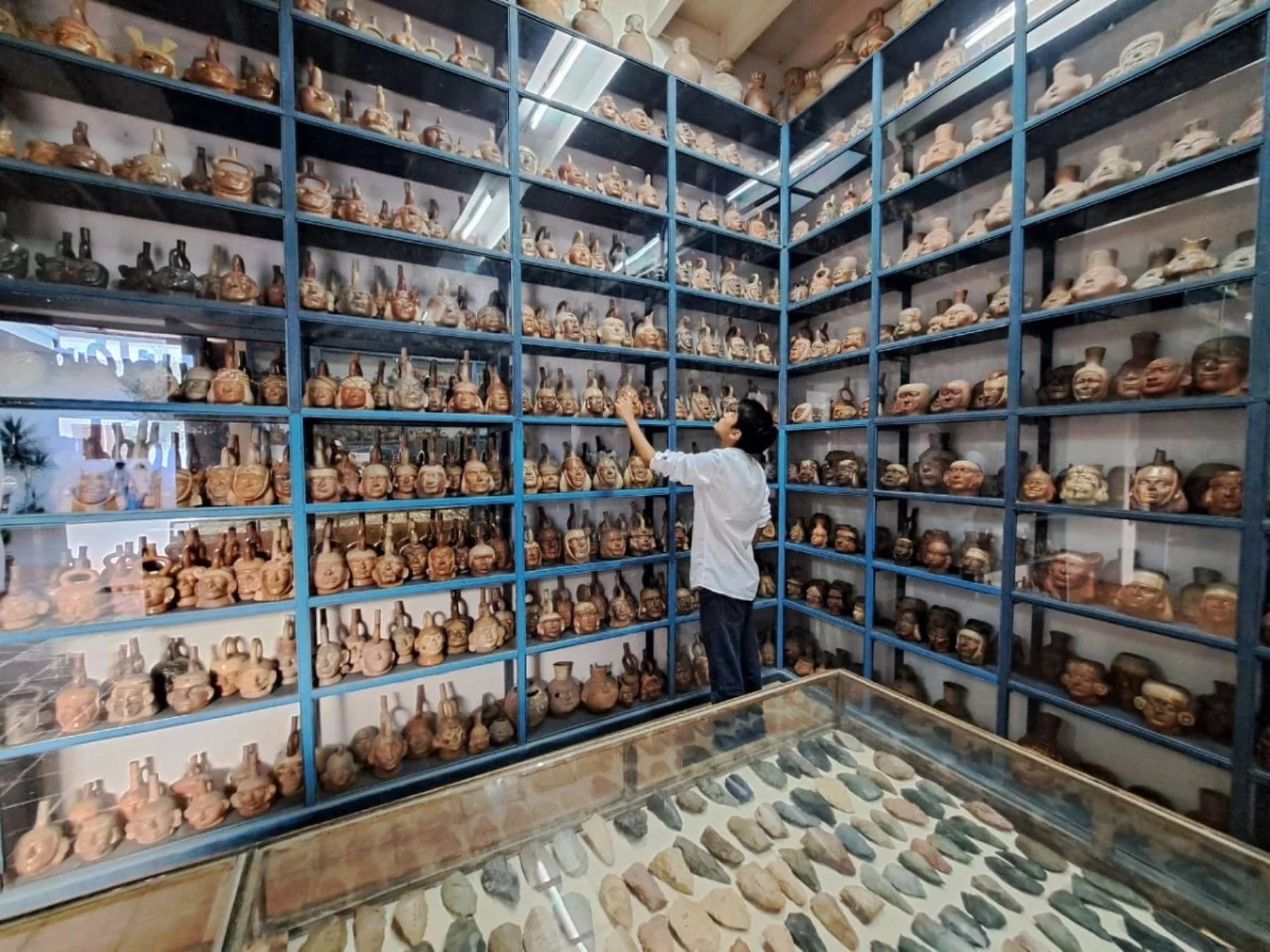 A person in a white shirt and dark pants browsing a large display of pottery artifacts in a museum or gallery. The artifacts are arranged on blue shelves and include various shaped ceramic vessels and figurines, with additional stones displayed in a glass case in front.