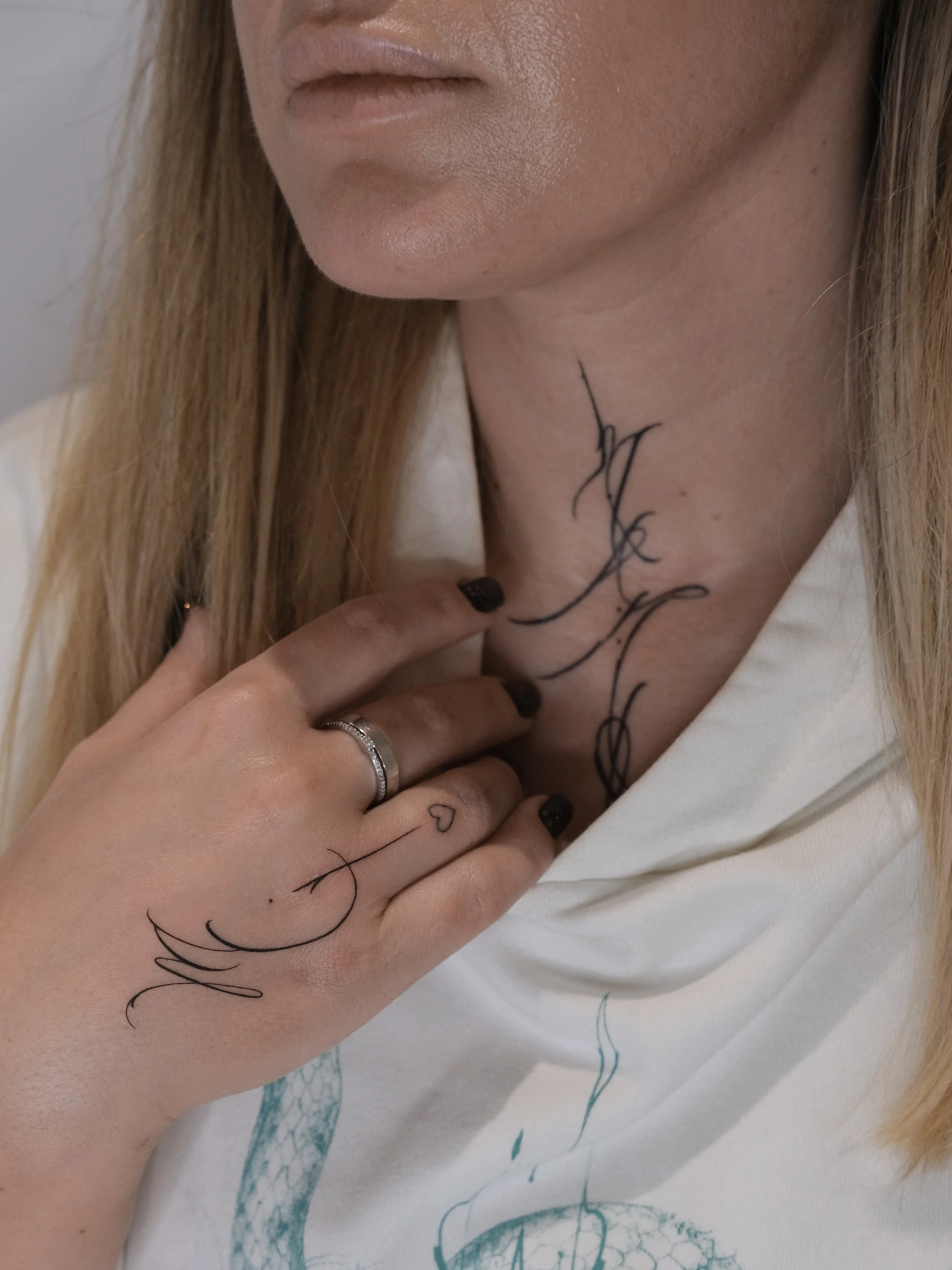 Close-up of a woman's neck and hand, showing a black script tattoo on her neck and hand, with her fingers touching the tattoo.