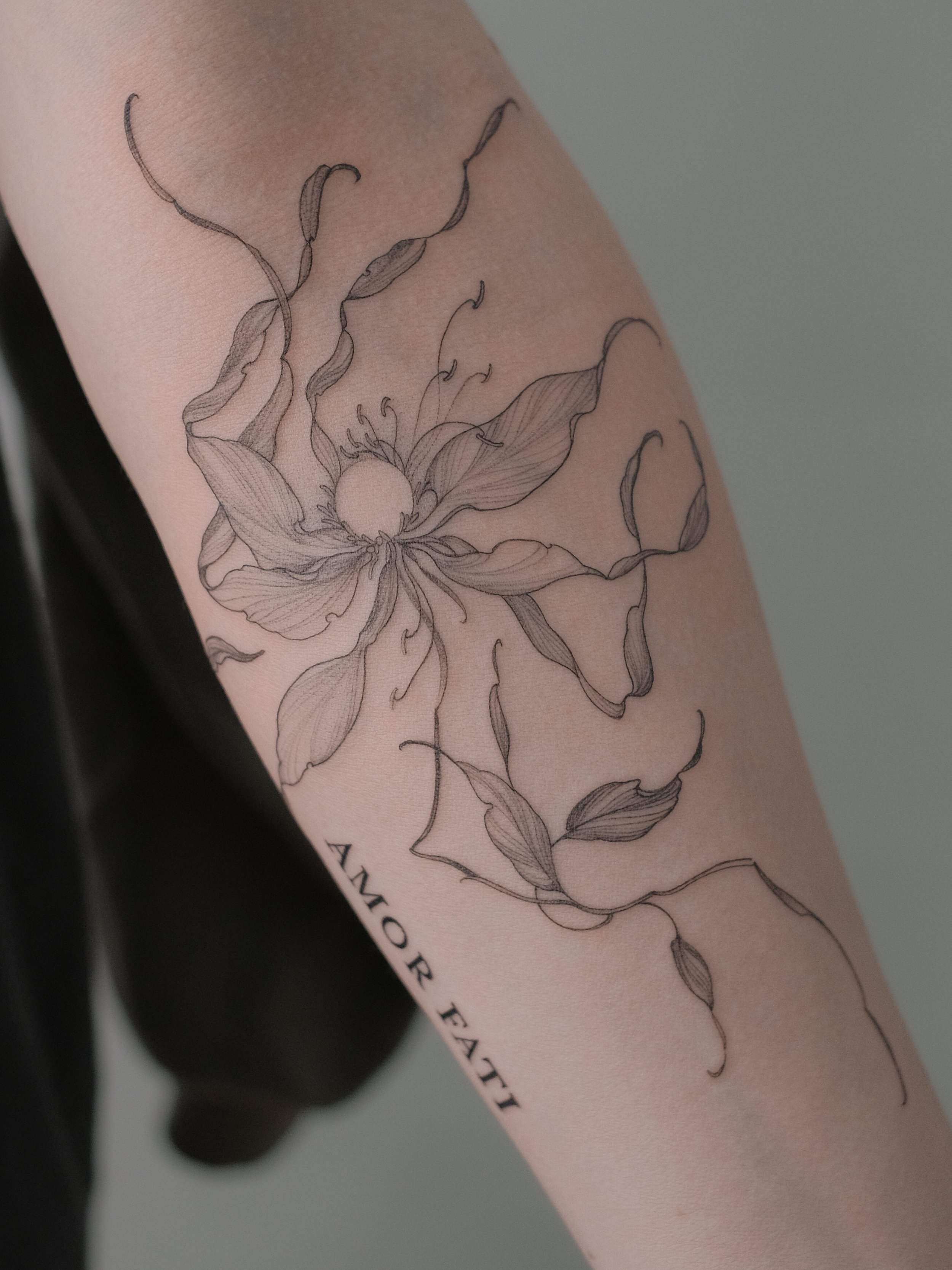 A tattoo of a detailed line drawing of a flower on a person's arm, with some text partially visible underneath it.