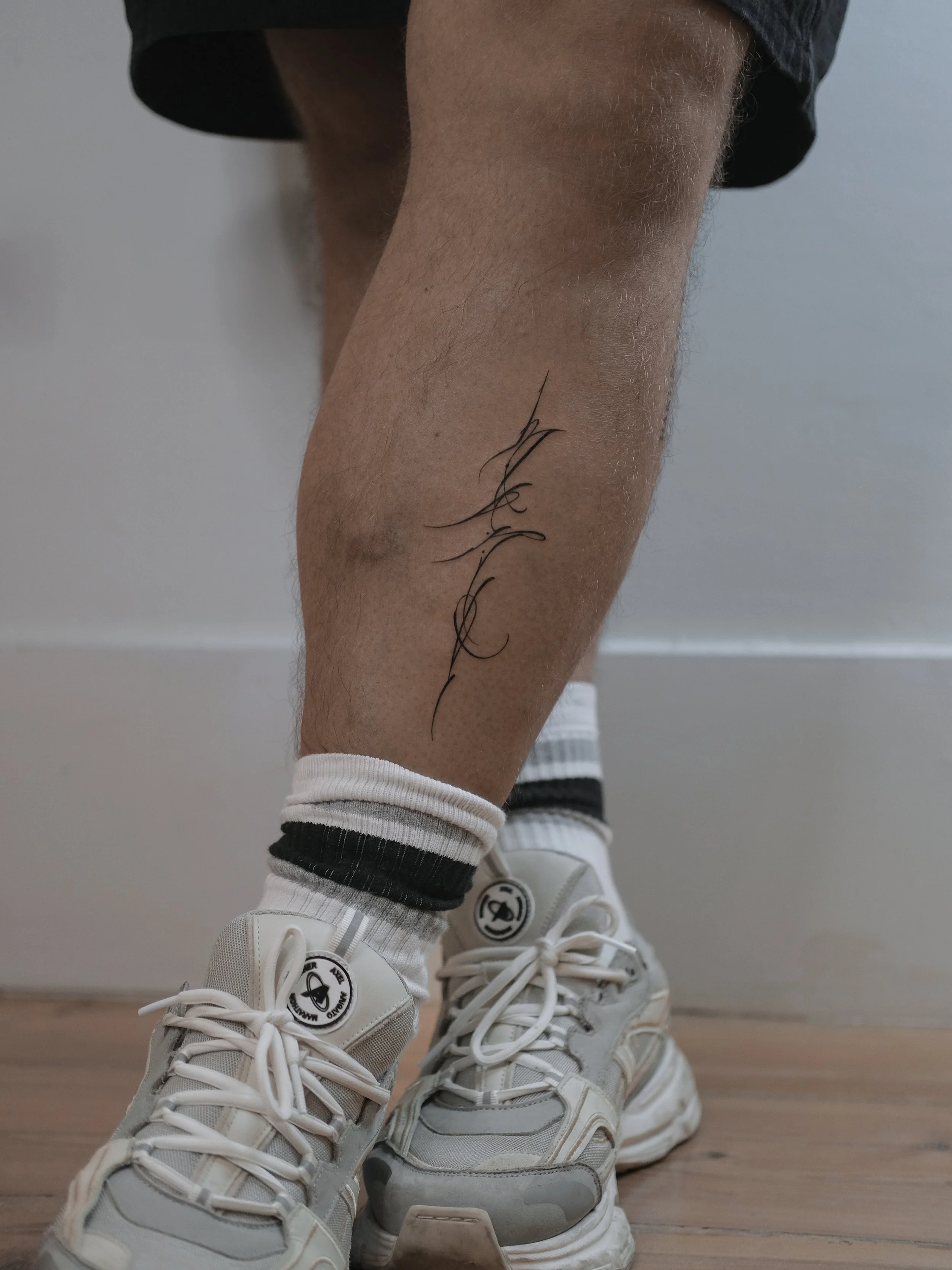 A person's lower leg with a tattoo of the word 'Love' in cursive script. The person is wearing white sneakers, striped socks, and black shorts.