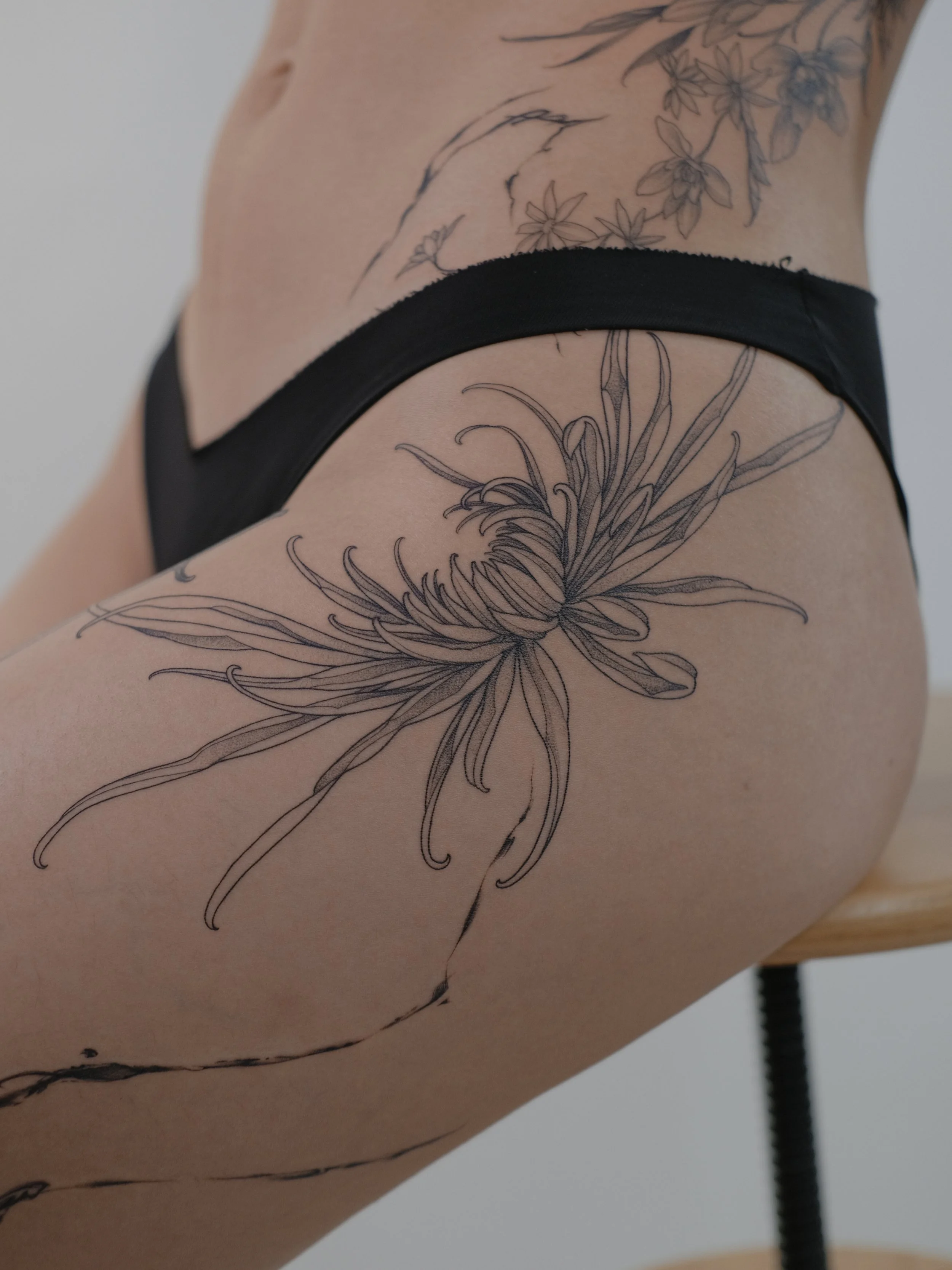 Close-up of a person's side and back, showing detailed black line floral tattoos on their skin, wearing black underwear.