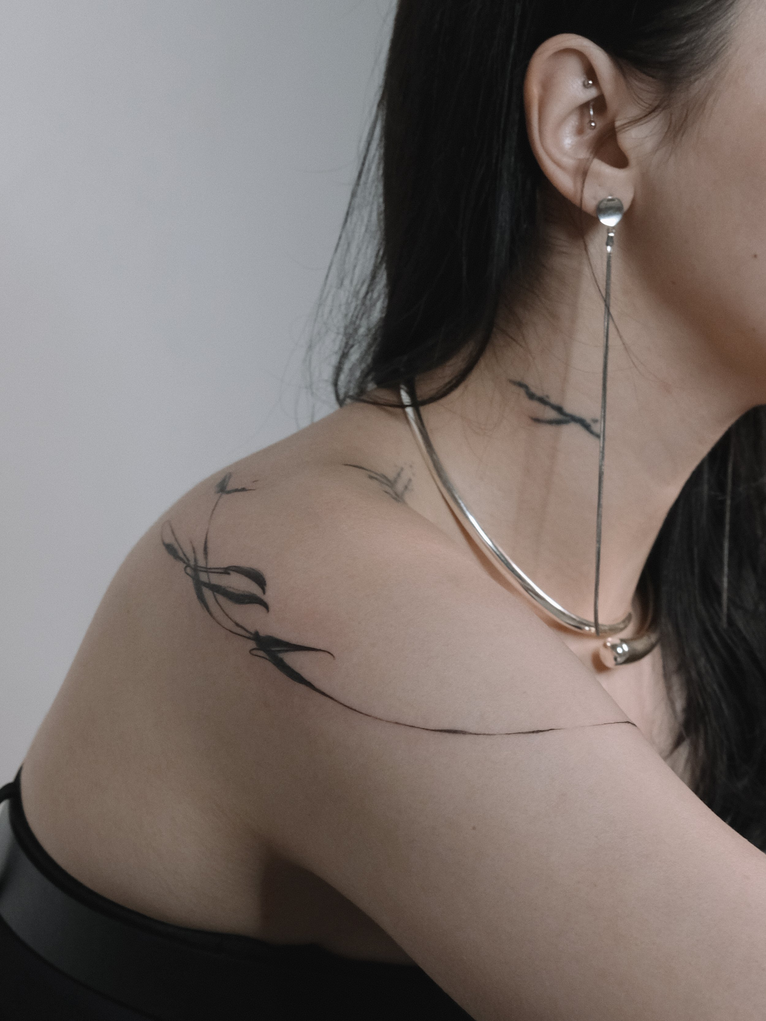 Close-up of a woman's shoulder and neck, showing a tattoo of a branch with leaves, and various earrings in her ear.