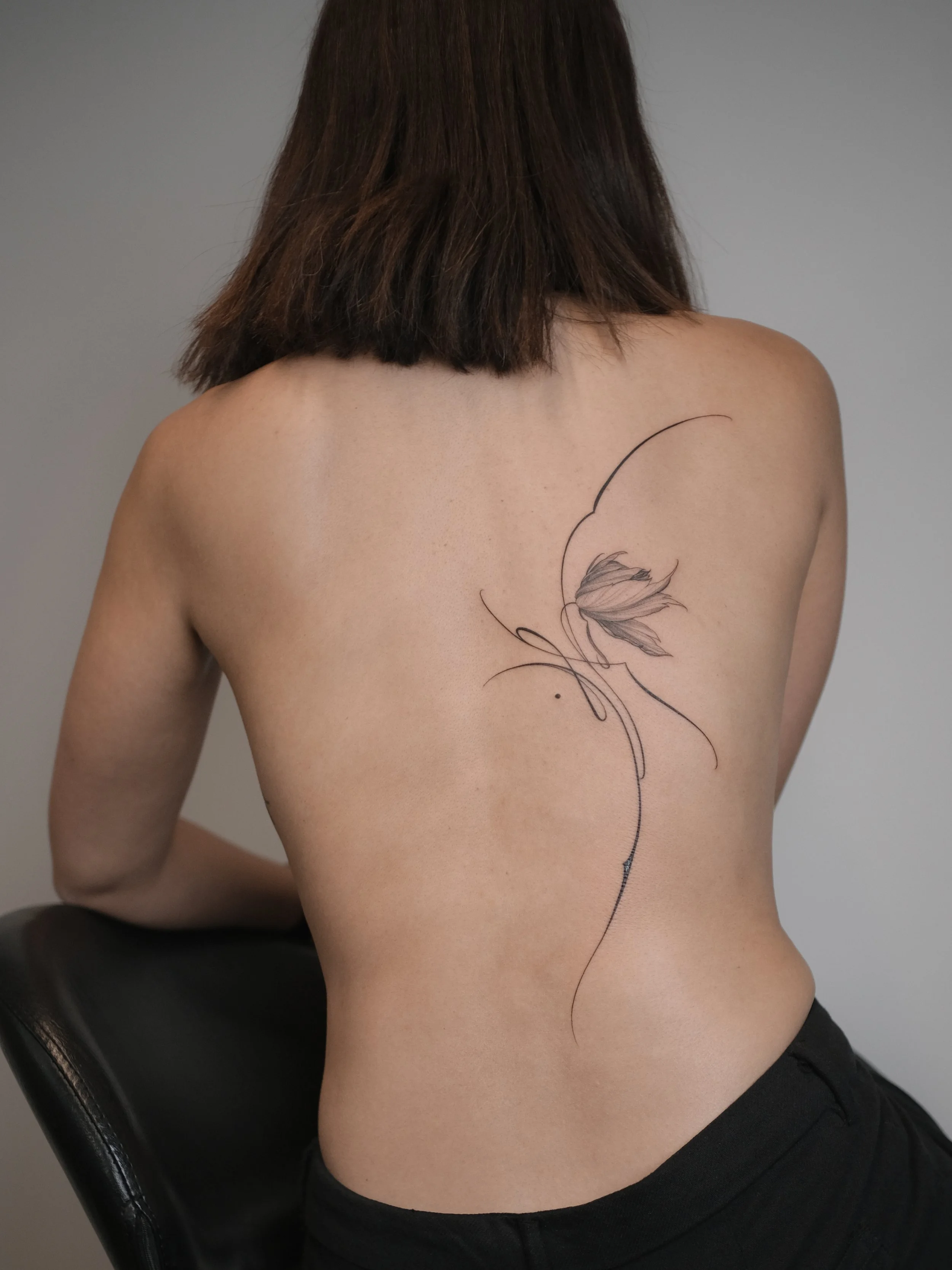 Back view of a woman with shoulder-length brown hair, sitting on a chair, and showing a floral tattoo on her upper back.