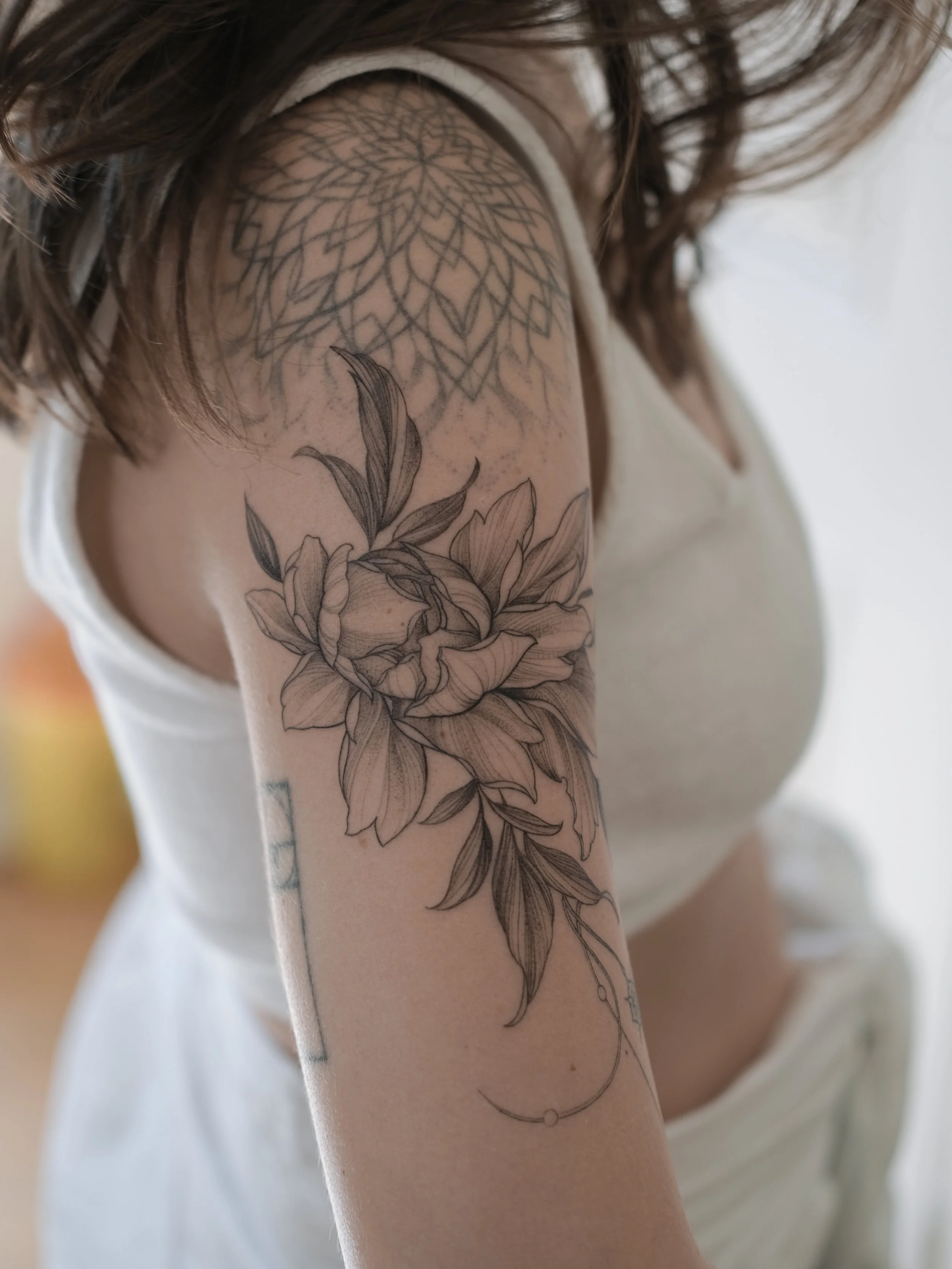 Close-up of a person's arm with a detailed black and gray floral tattoo, including a large flower and leaves, and a geometric pattern tattooing the upper arm and shoulder.