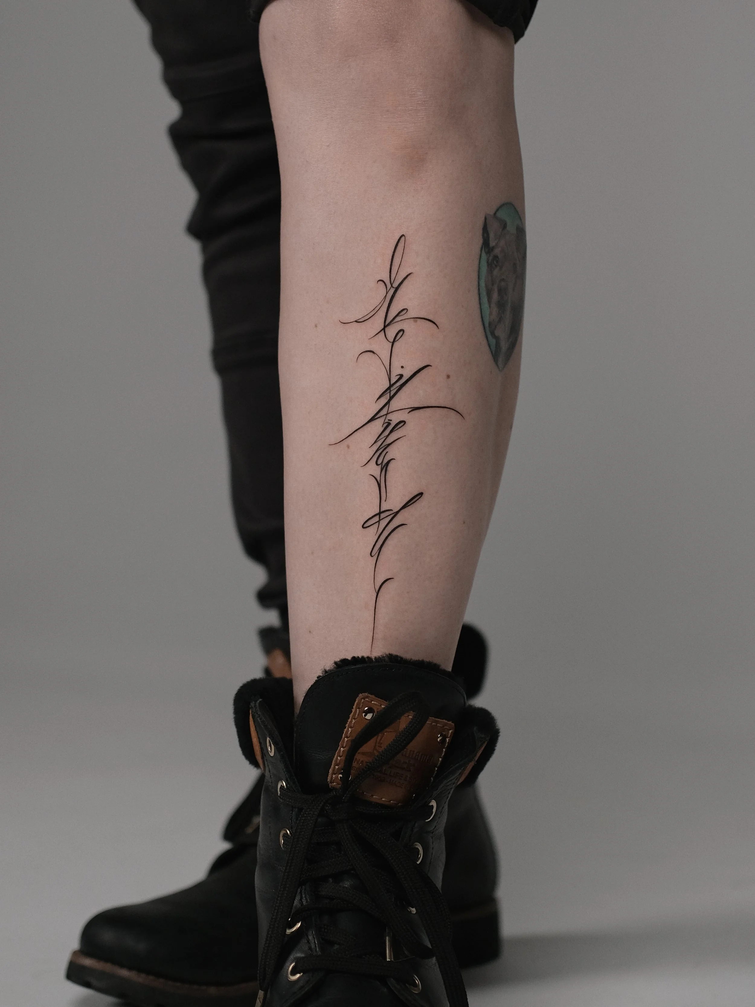 Close-up of a person's leg with a tattoo of a dog's face inside a blue circle and a vertical script tattoo on the calf, wearing black pants and black boots.