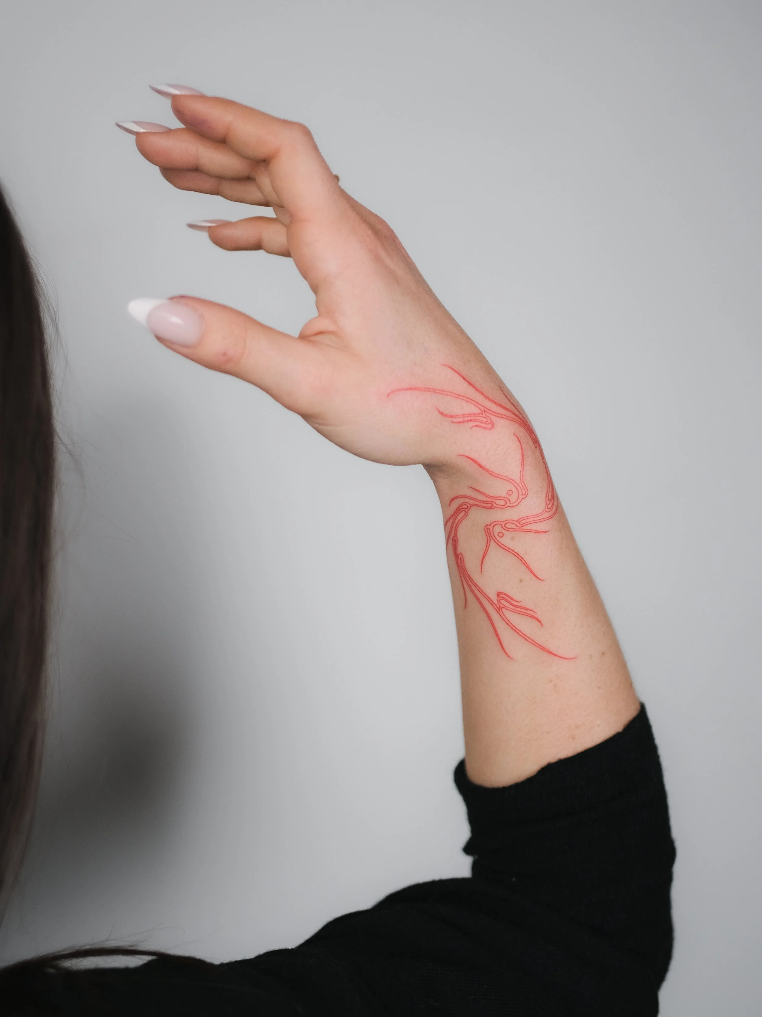 A person's arm raised with a red anatomical tattoo of the veins and arteries on the inner side of the wrist and forearm, wearing a black long sleeve shirt, with a plain light gray wall in the background.