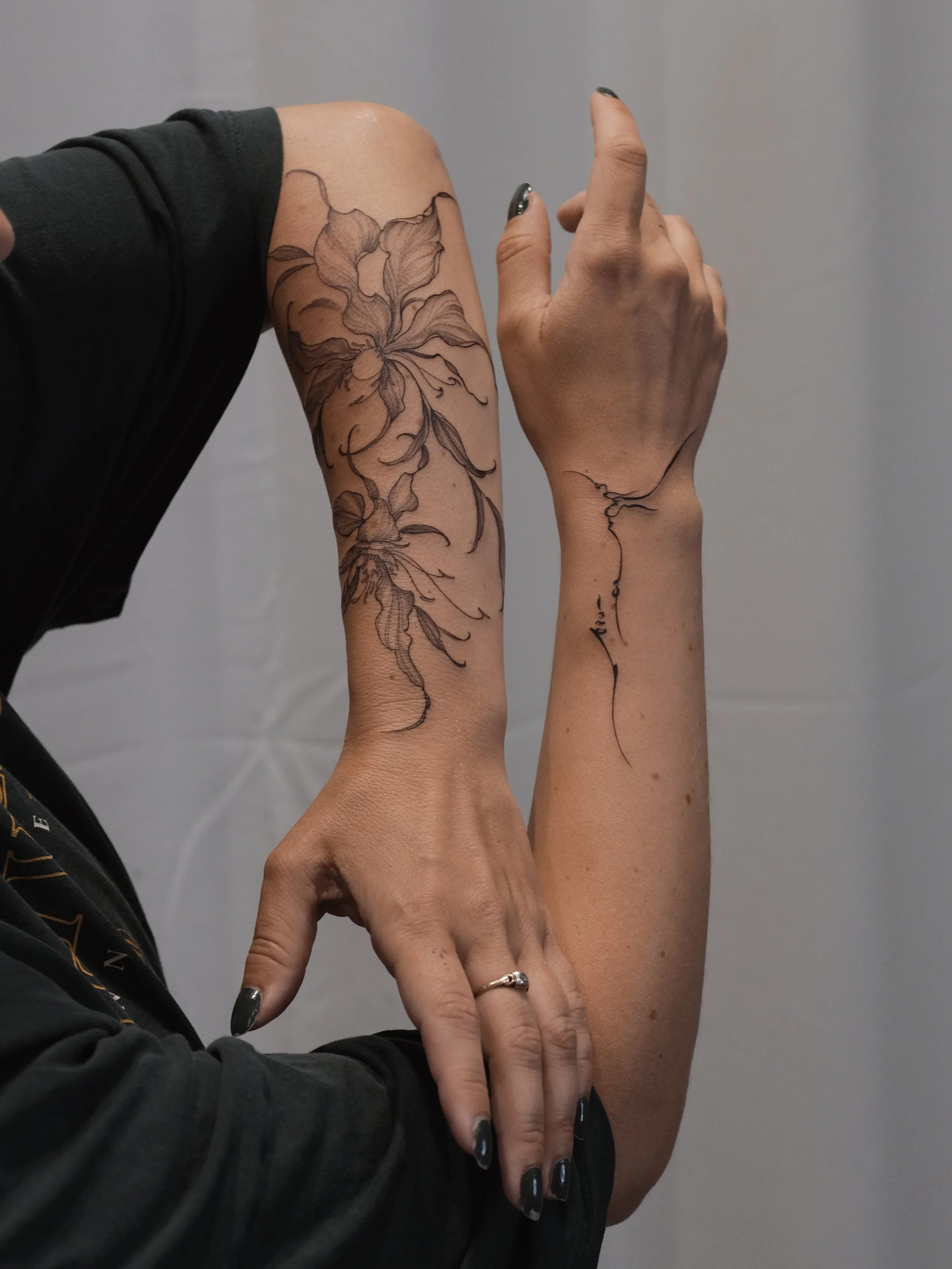 Close-up of two arms, one with a floral tattoo and the other with a fine line tattoo, against a plain gray background.