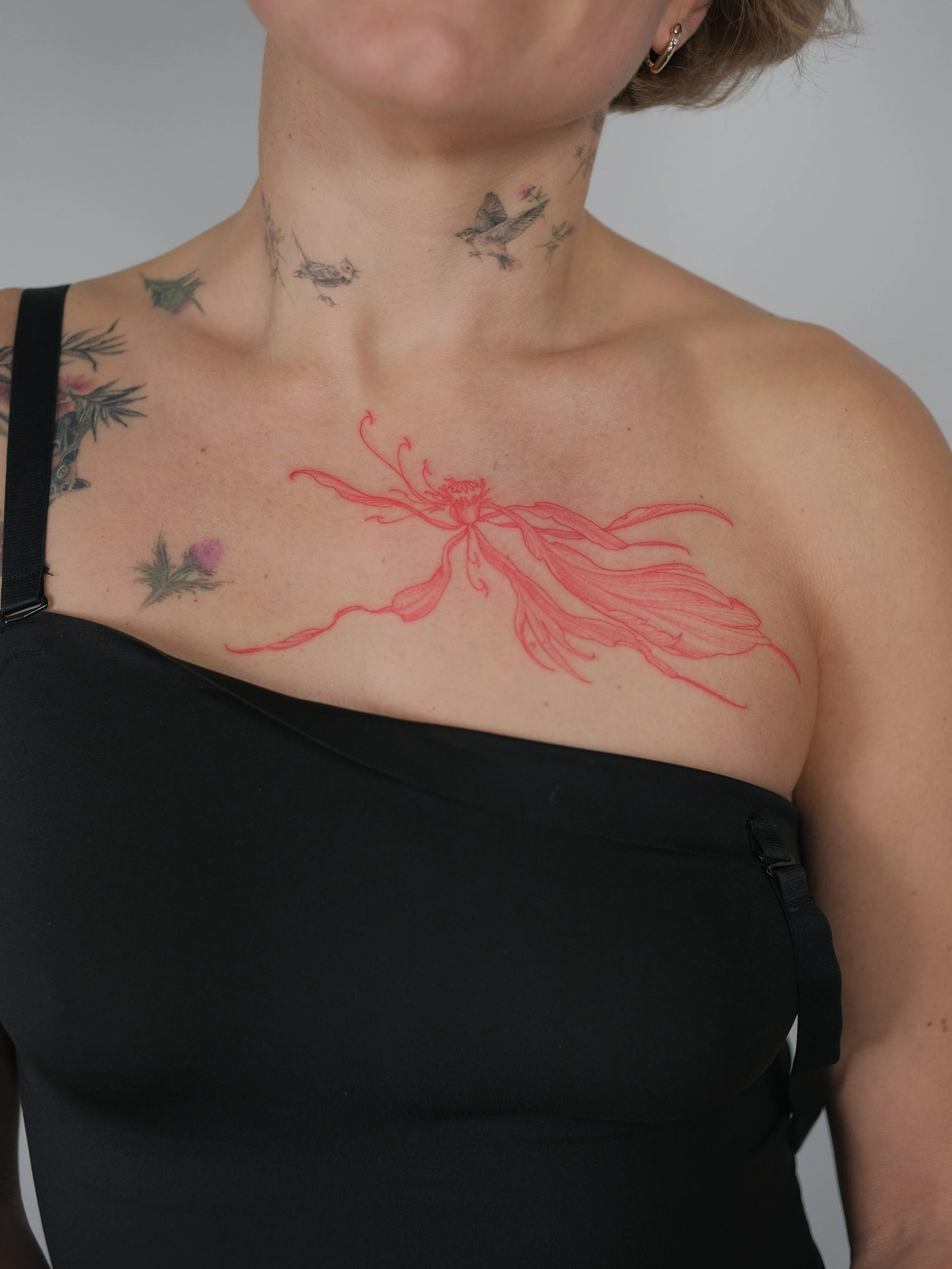 Close-up of a woman's upper chest and neck showing tattoos, including a large pink flower on the chest, small birds on the neck, and other smaller designs on the shoulder and neck area.