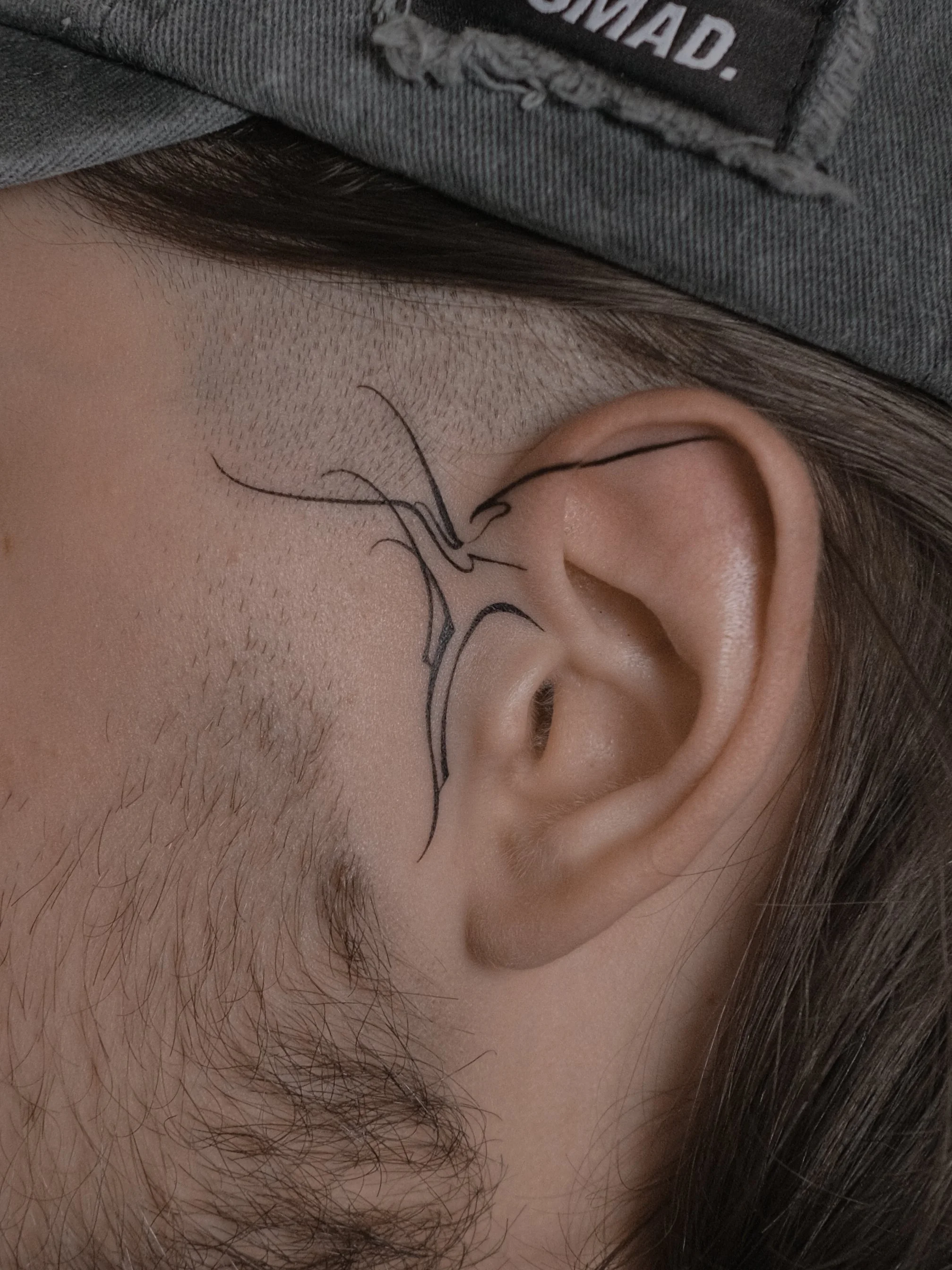 Close-up of a person's ear, along with facial hair and a tattoo on the cheek, with a gray shirt and part of a black label visible.