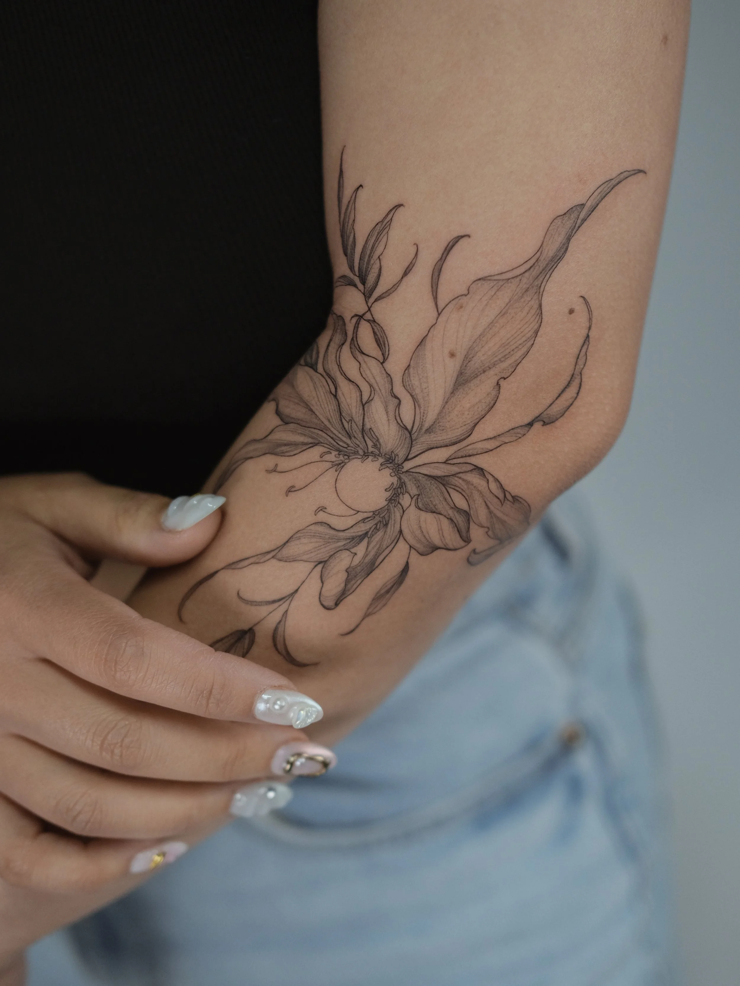 A close-up of a person's forearm with a black ink floral tattoo design, featuring large leaves and detailed flower petals, with a portion of their hand showing white nail polish with pearl accents.