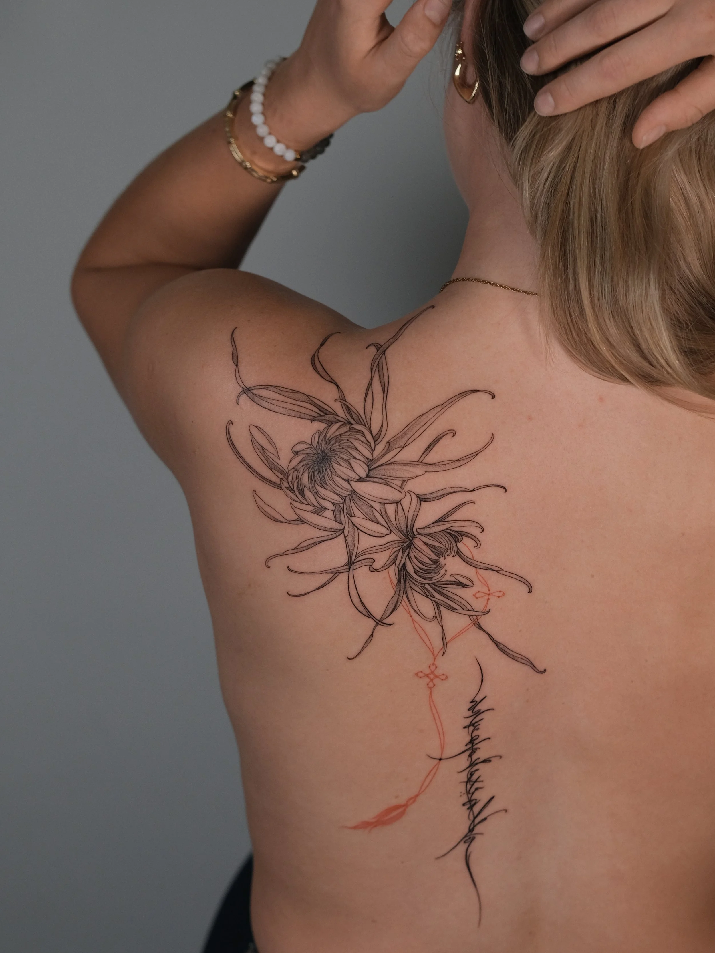 A woman with long, light-colored hair posing with her back to the camera. She has a large floral tattoo on her left shoulder blade, and is wearing gold jewelry including earrings, necklaces, and bracelets. She is holding her hair back with her right 