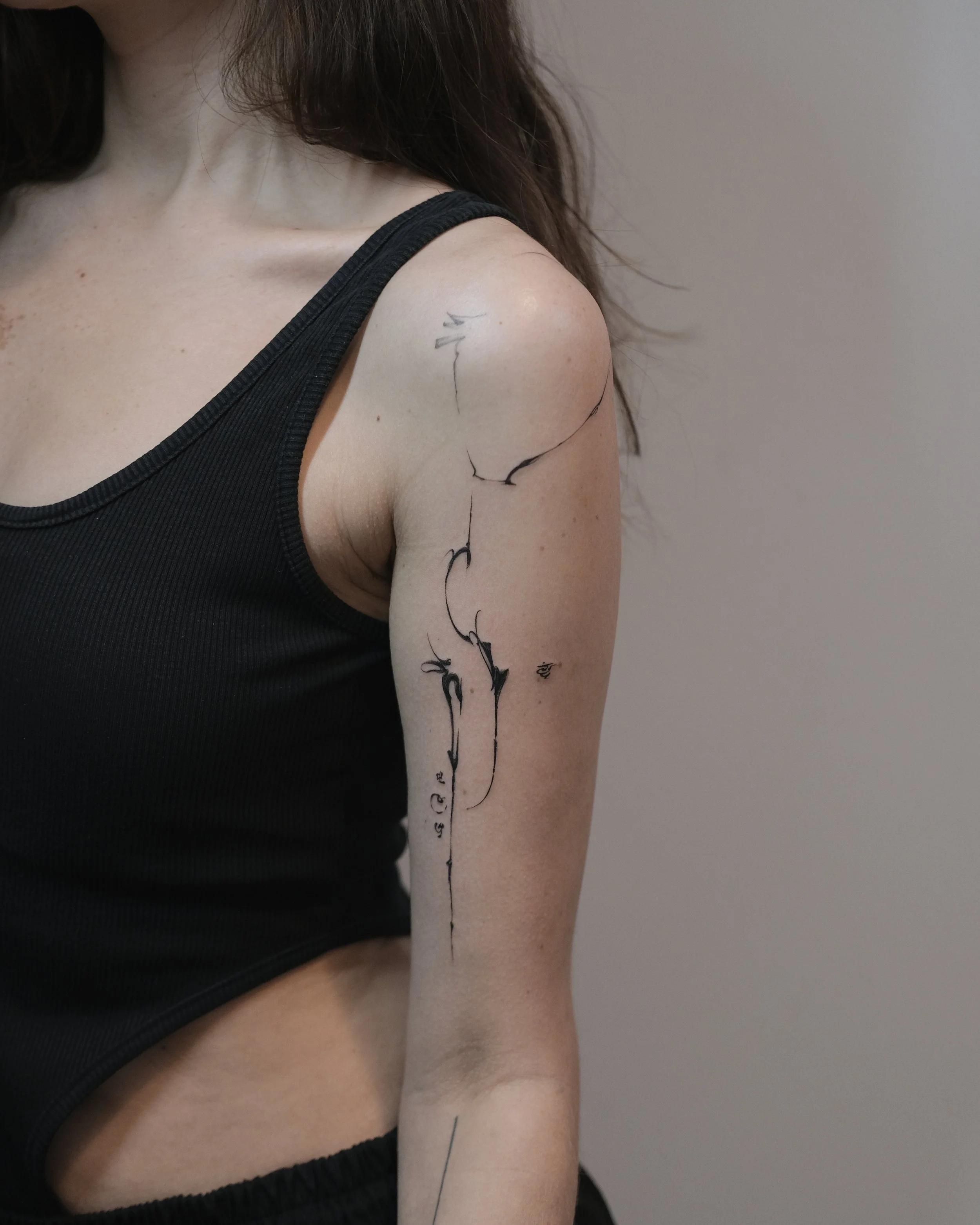 Close-up of a woman's shoulder and upper arm with black ink tattoo lines, wearing a black tank top, against a plain background.