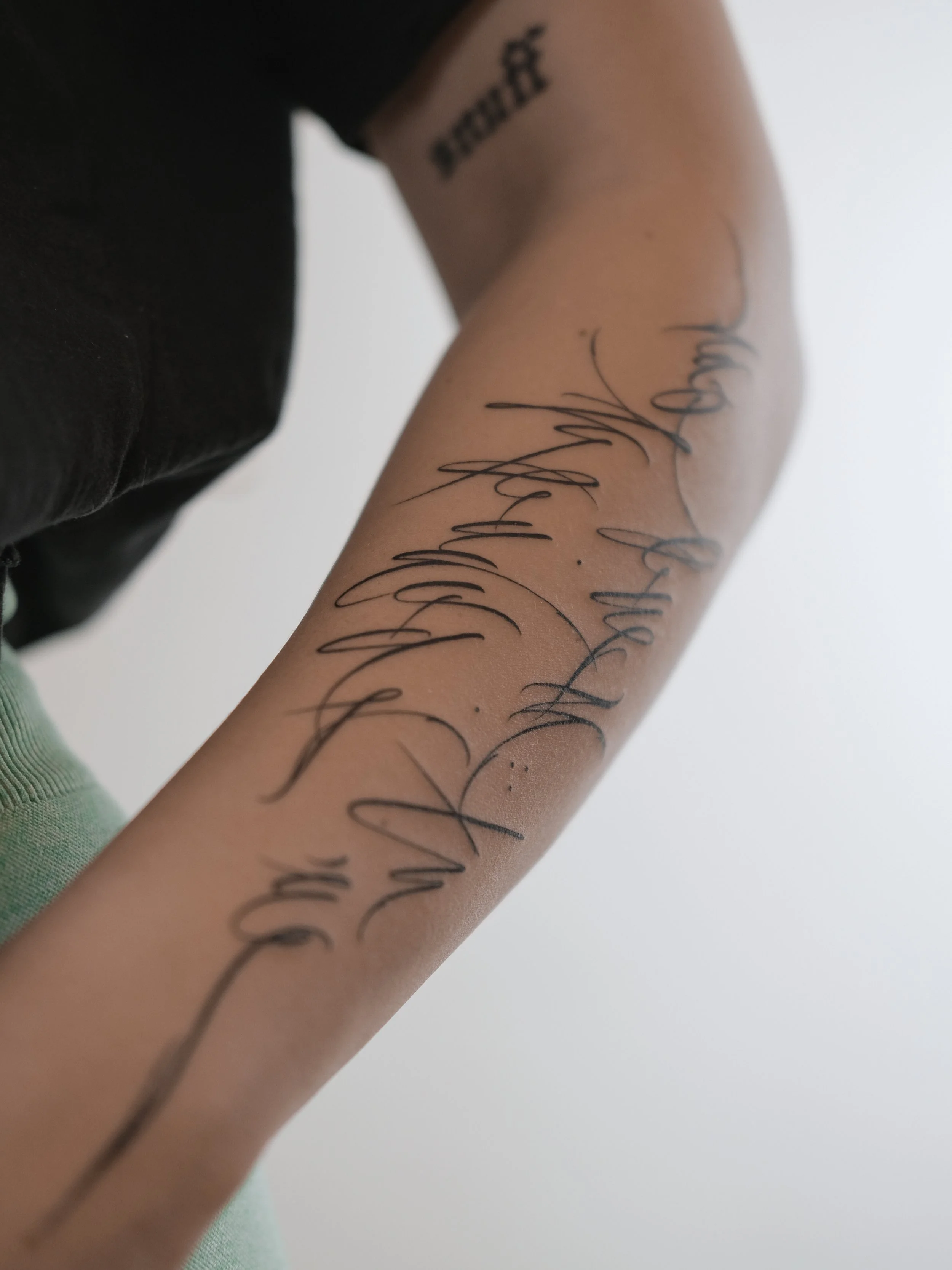Close-up of a person's arm with a tattoo of cursive writing, wearing a dark short sleeve shirt, against a plain background.