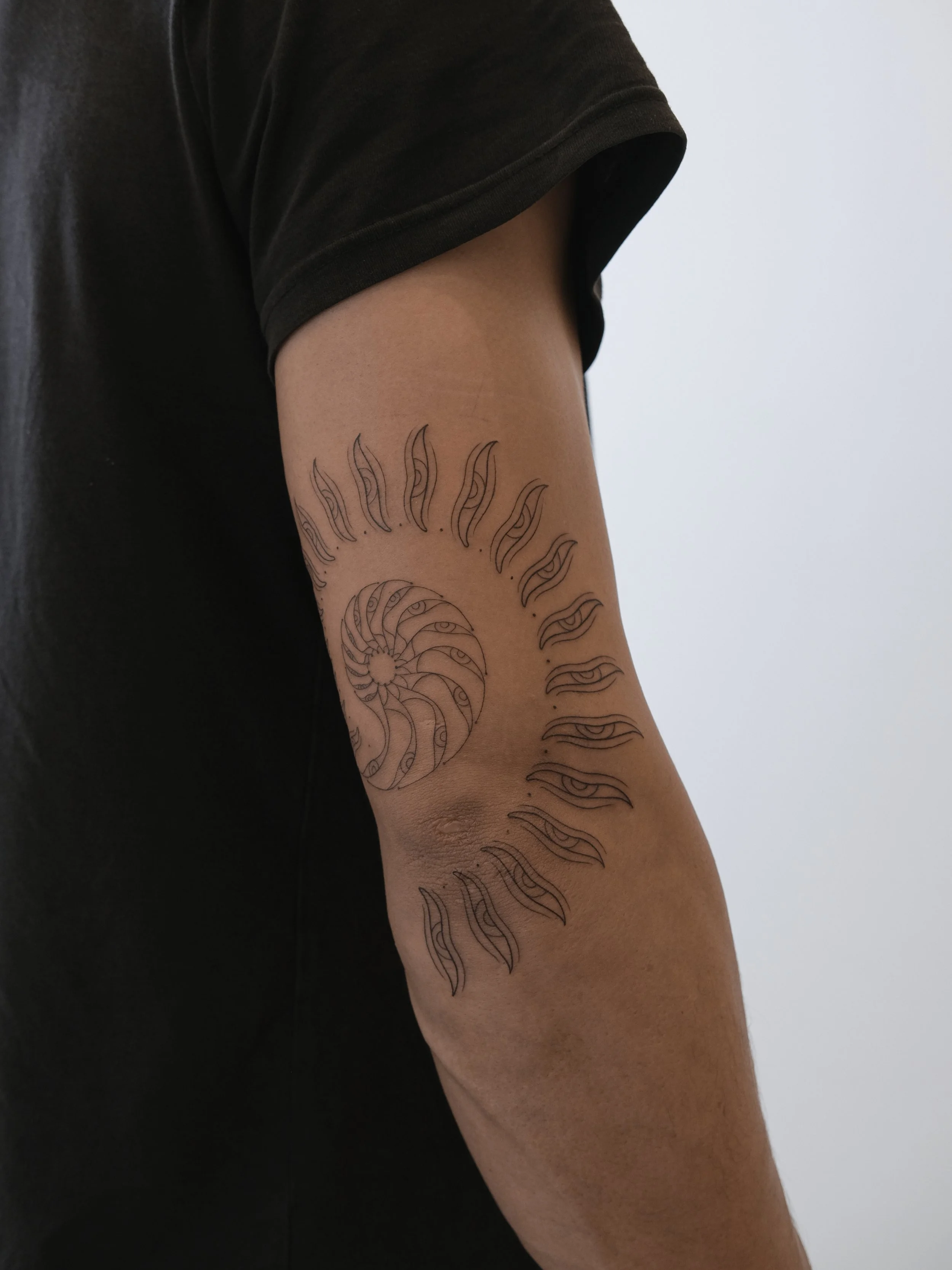 A person's arm with a tattoo of a spiral pattern and wavy eye designs, wearing a black short-sleeved shirt.