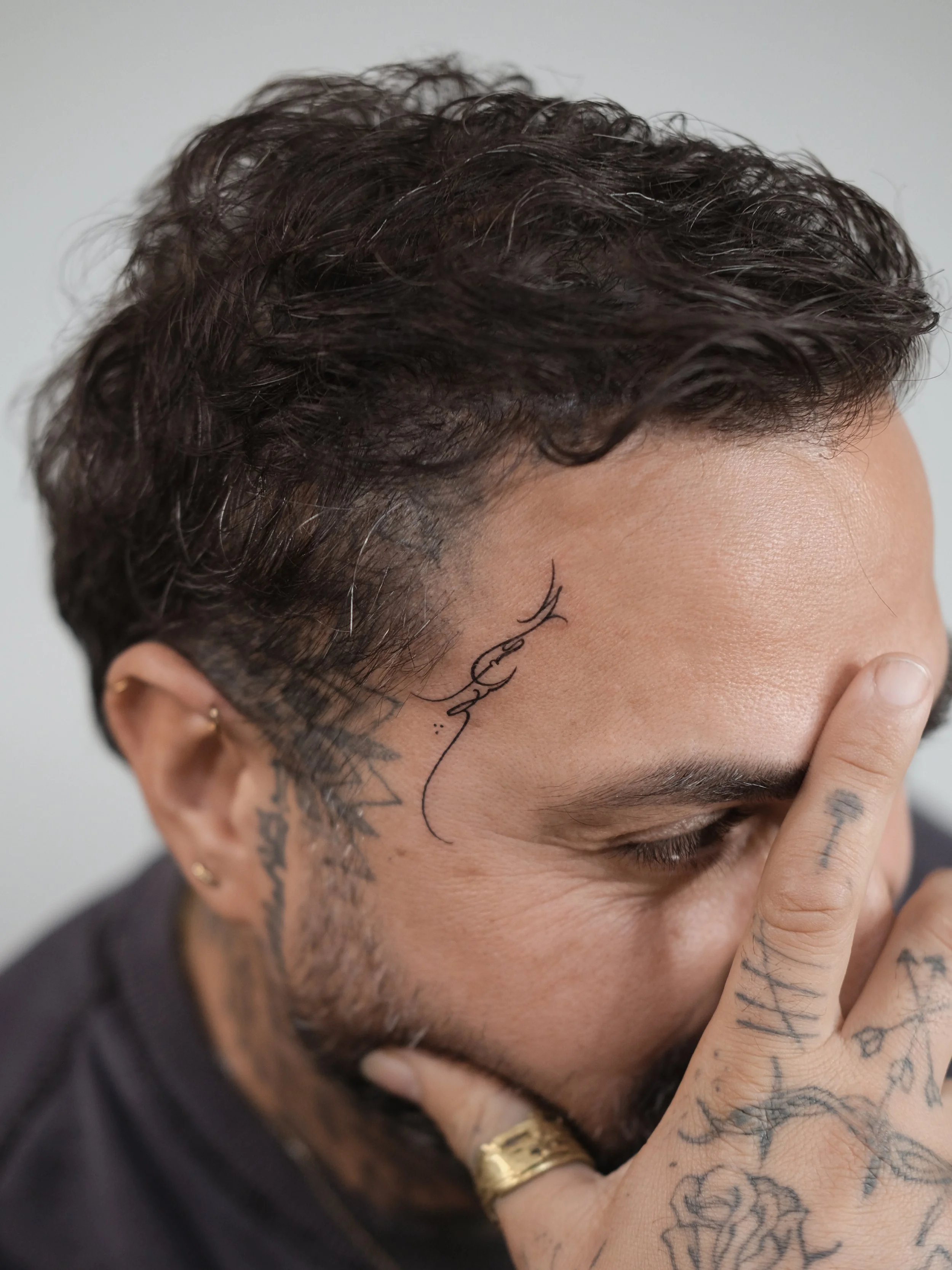 A person with short, curly dark hair covering part of their face, tattoos on their hand and face, and wearing multiple rings, with a hand resting on their face.