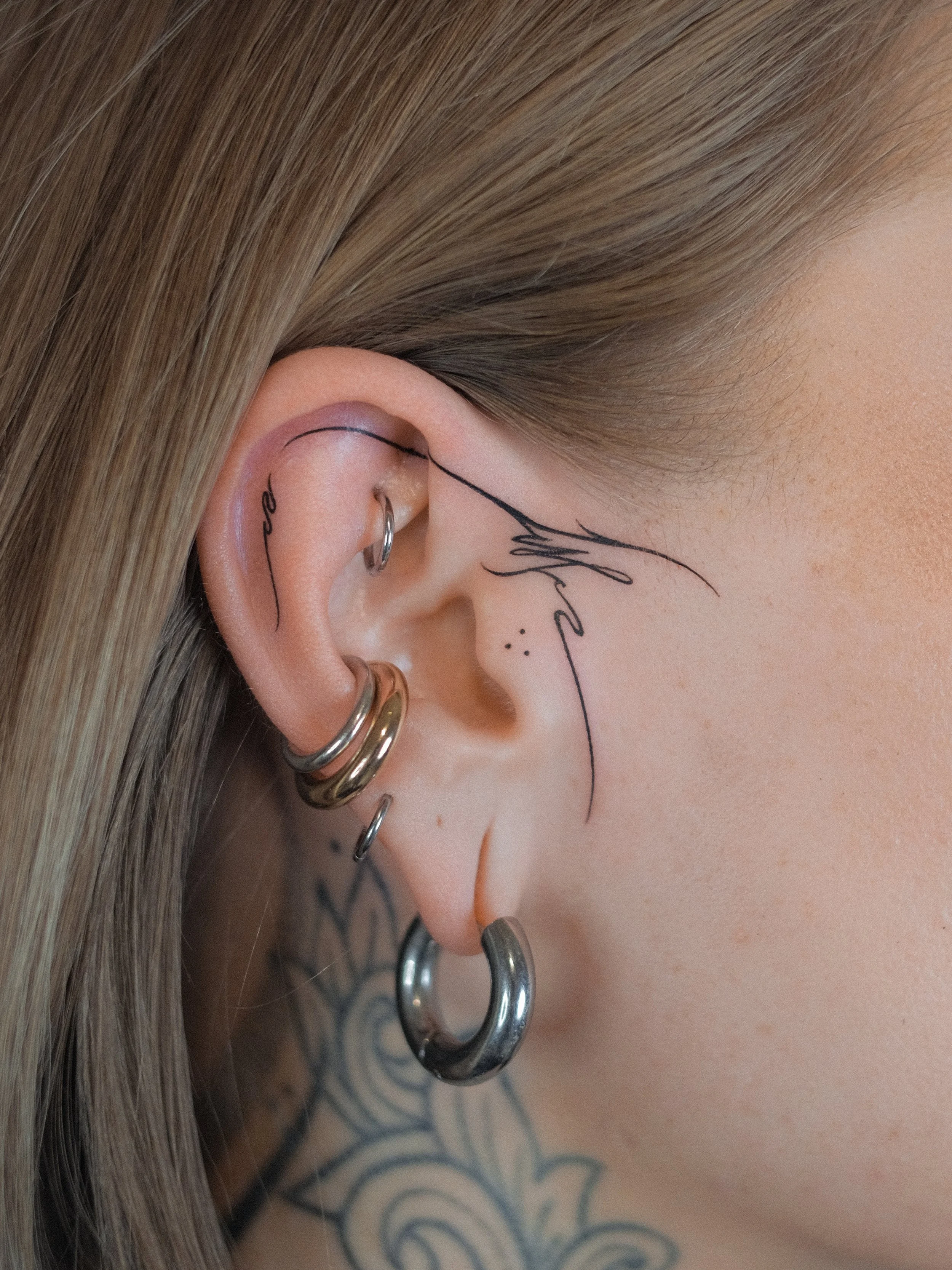 Close-up of a person's ear with multiple silver hoop earrings and a tattoo on the shoulder, with a tattooed line tracing the ear's outline.