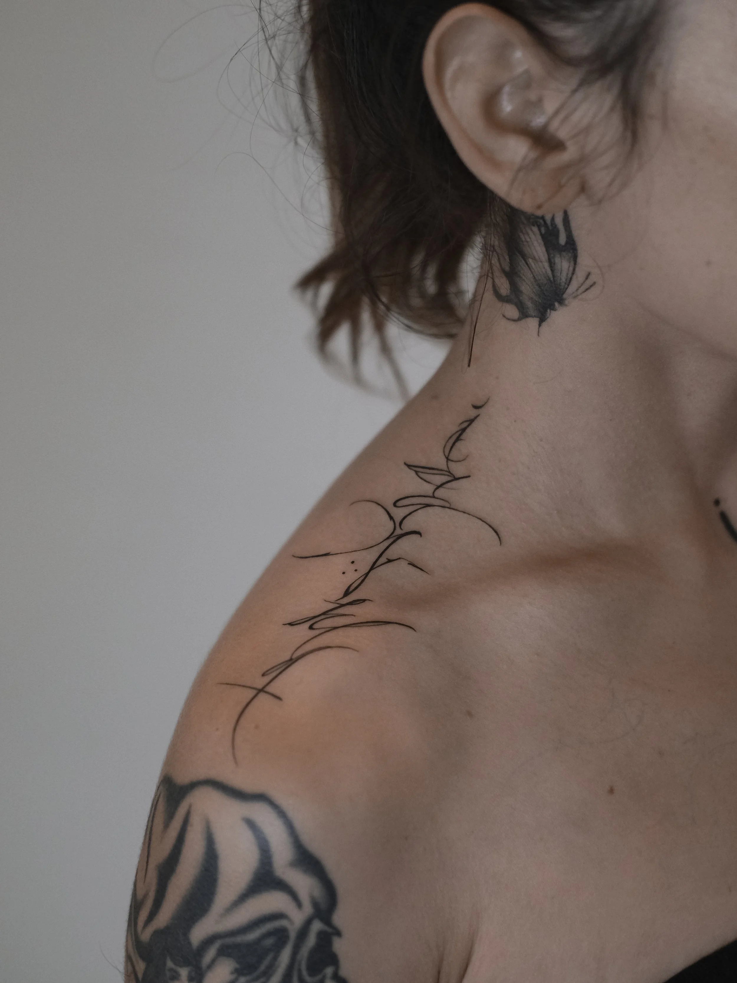 Close-up of a person's shoulder and neck with black tattoos, including a floral design and script, and a large earlobe with dark brown hair.