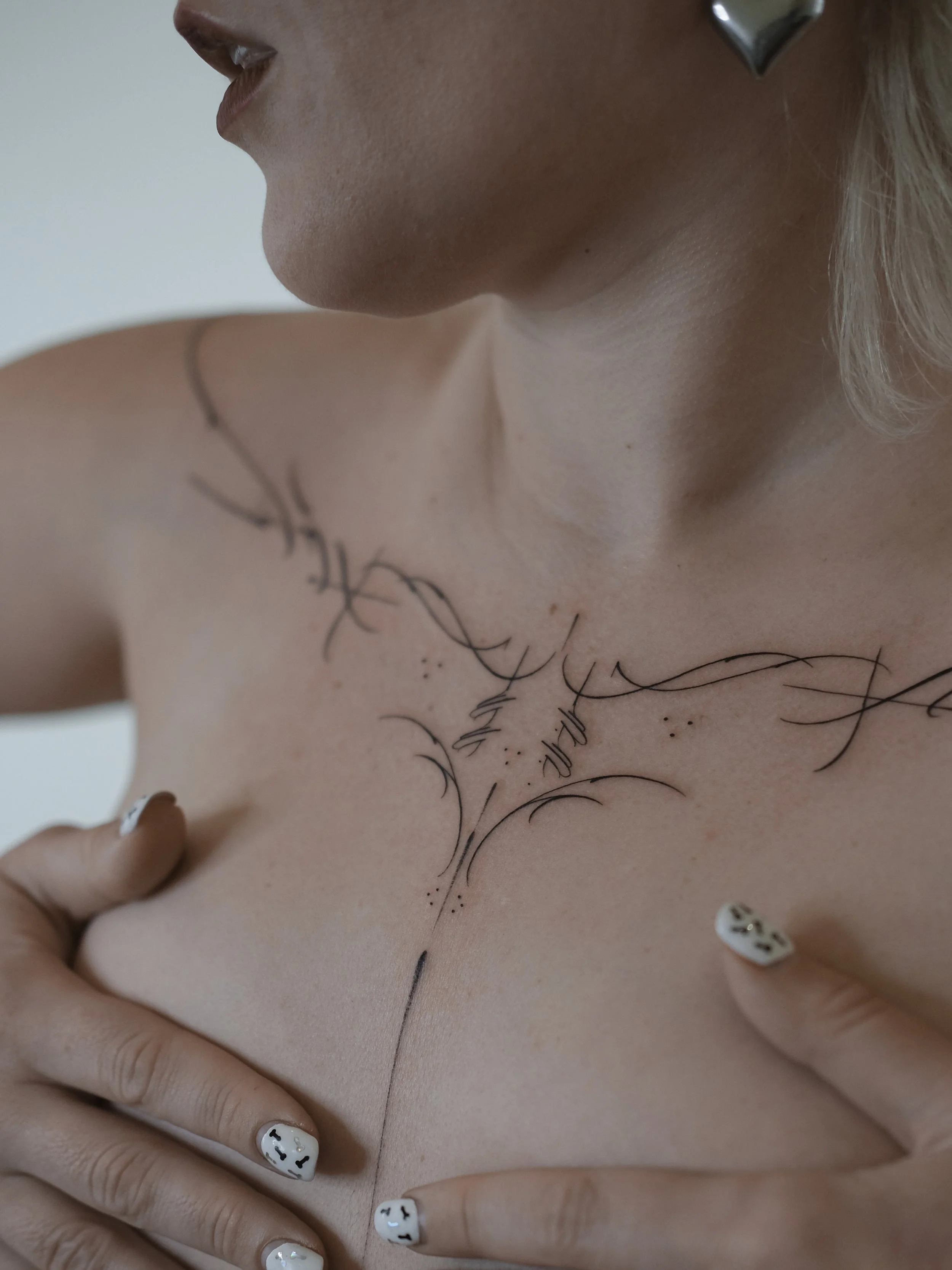 Close-up photo of a person's neck, shoulder, and chest, with black ink line art tattoo on the chest, and fingers with black-and-white nail art touching the skin.