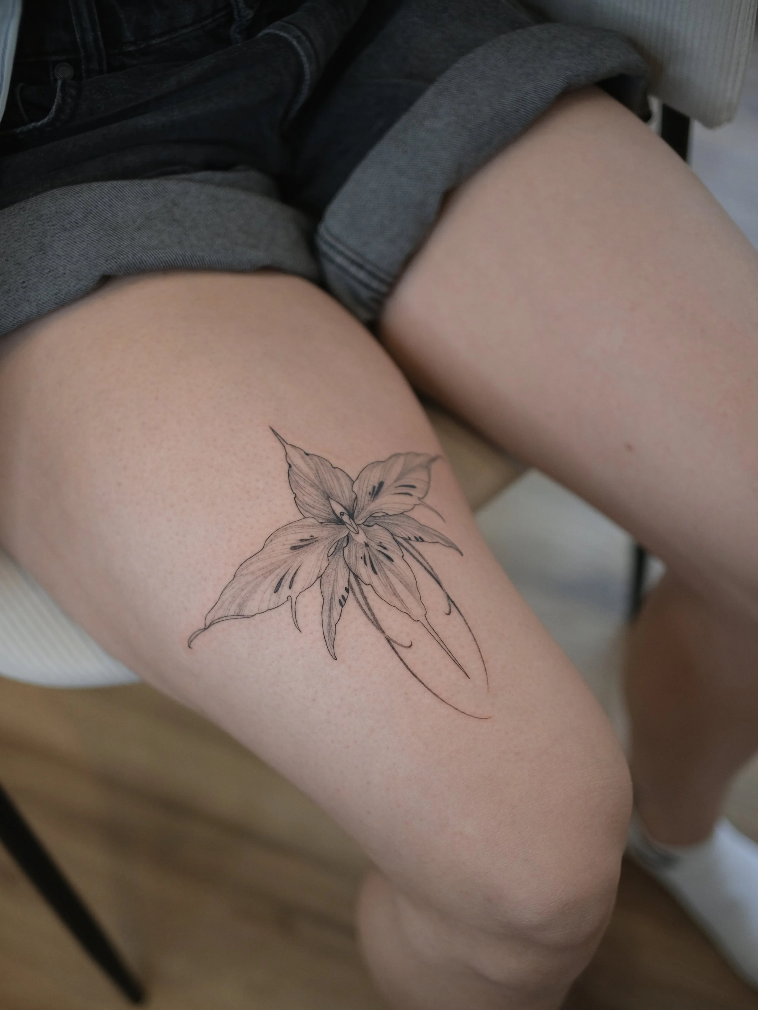 Close-up of a person's thighs with a black outline floral tattoo on the left thigh and wearing gray denim shorts.