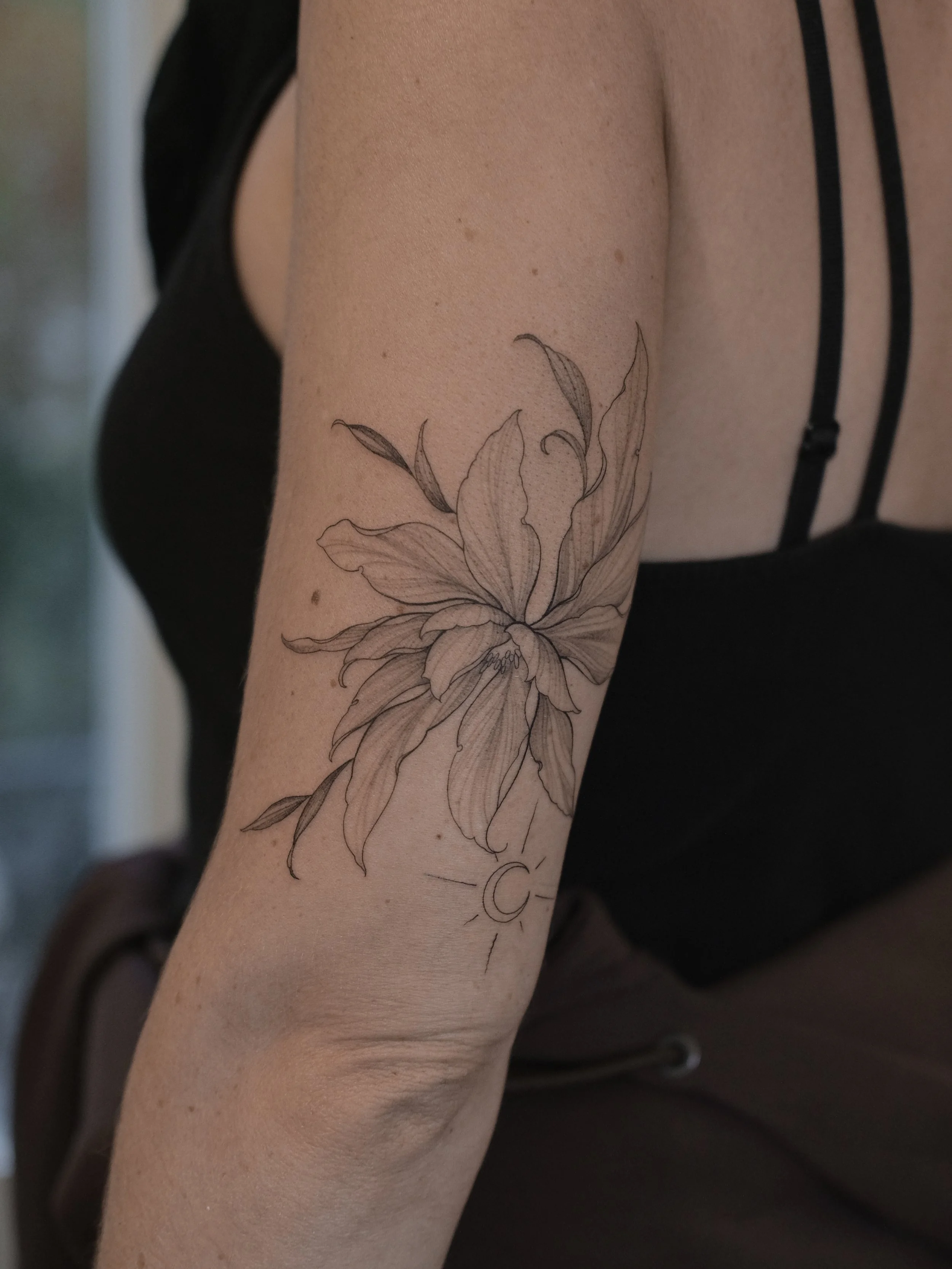 A close-up of a person's arm featuring a detailed black and gray flower tattoo. The person is wearing a black strap top.