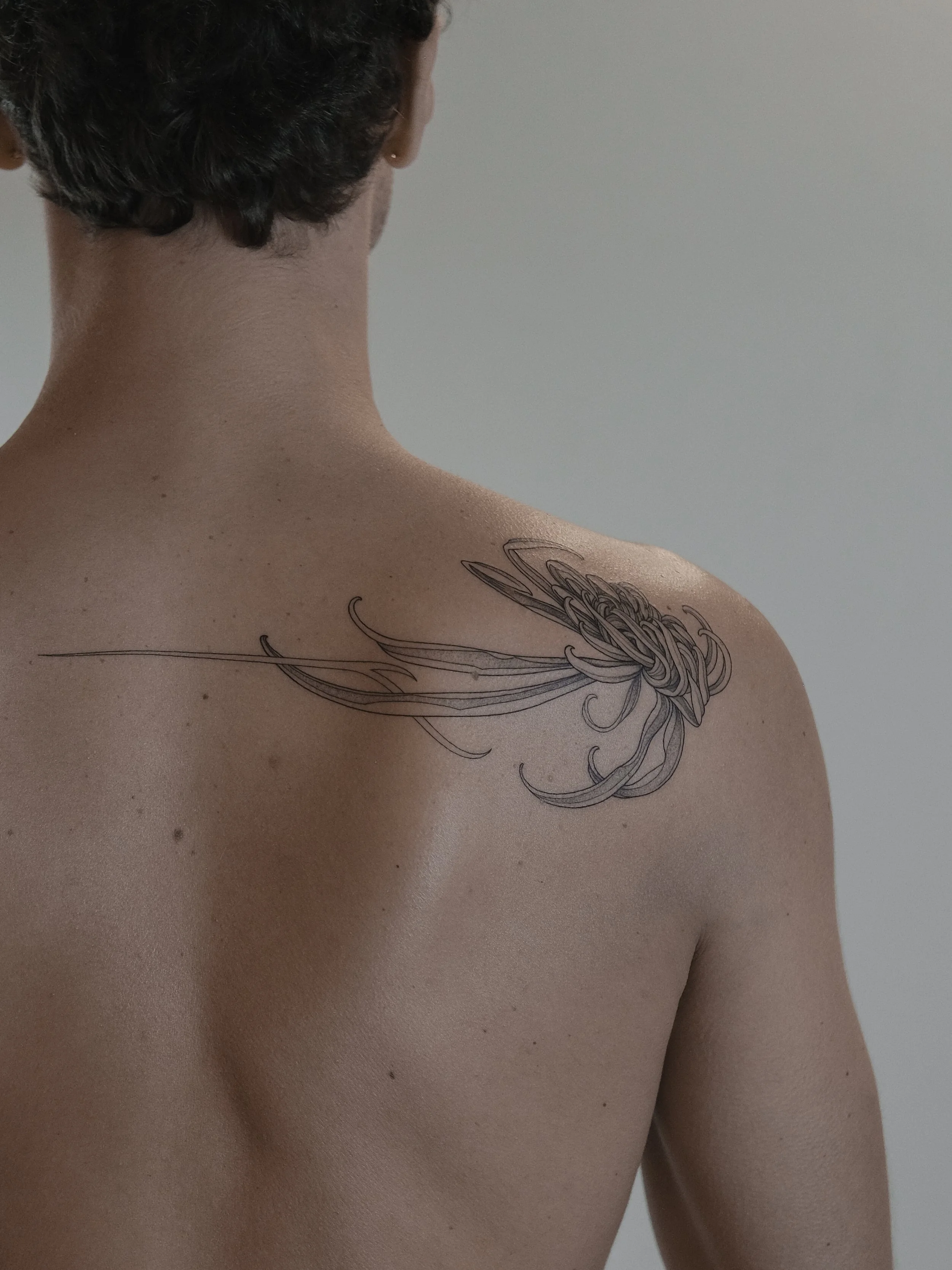 A person with short dark hair and light skin, viewed from behind and to the side, showing their back, neck, shoulder, and a detailed black and gray line drawing tattoo of a squid on their shoulder.