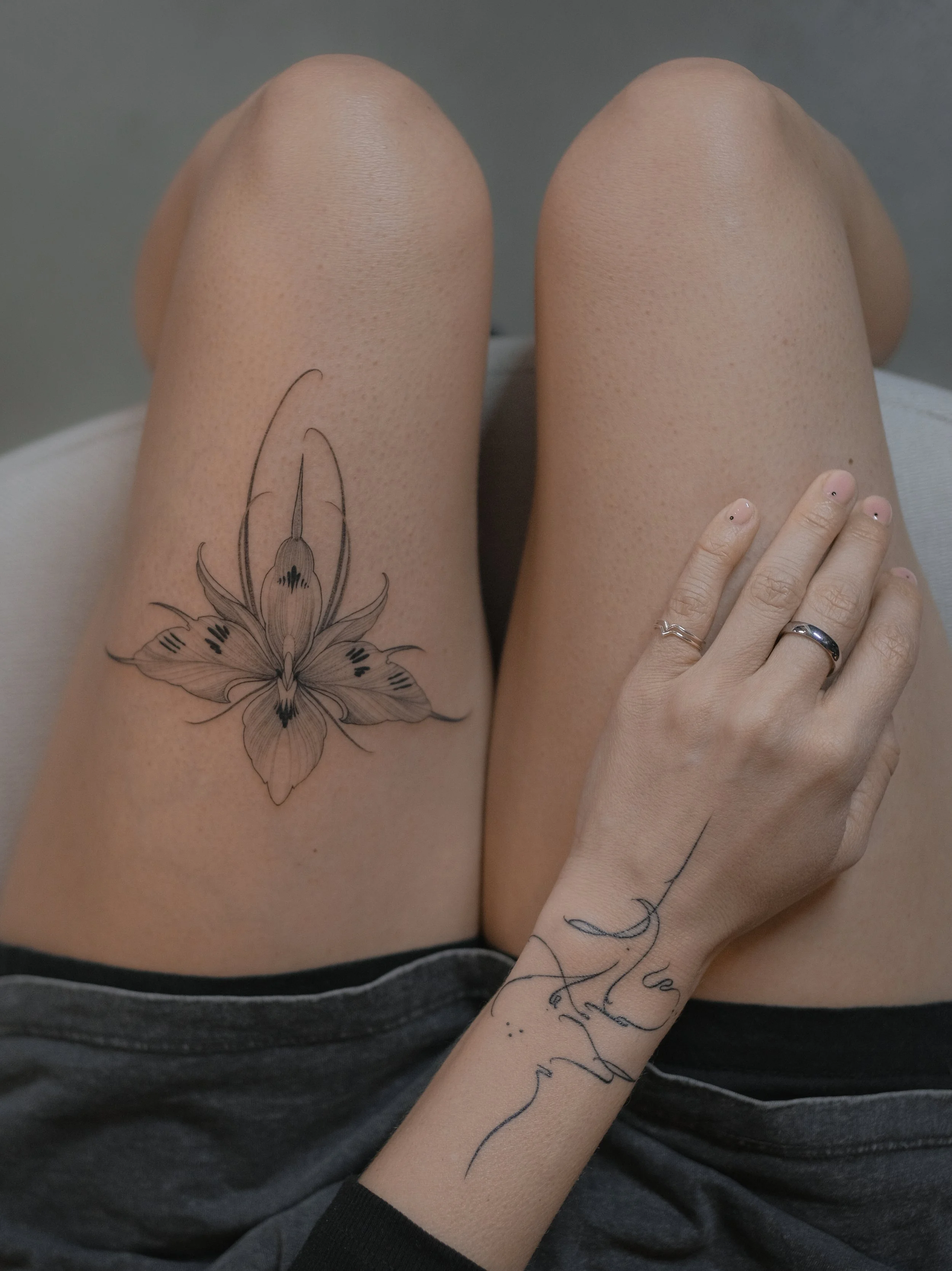 Close-up of a person's thigh with a detailed lily flower tattoo and a tattooed arm resting on their leg. The person is wearing dark gray shorts and the background is neutral gray.