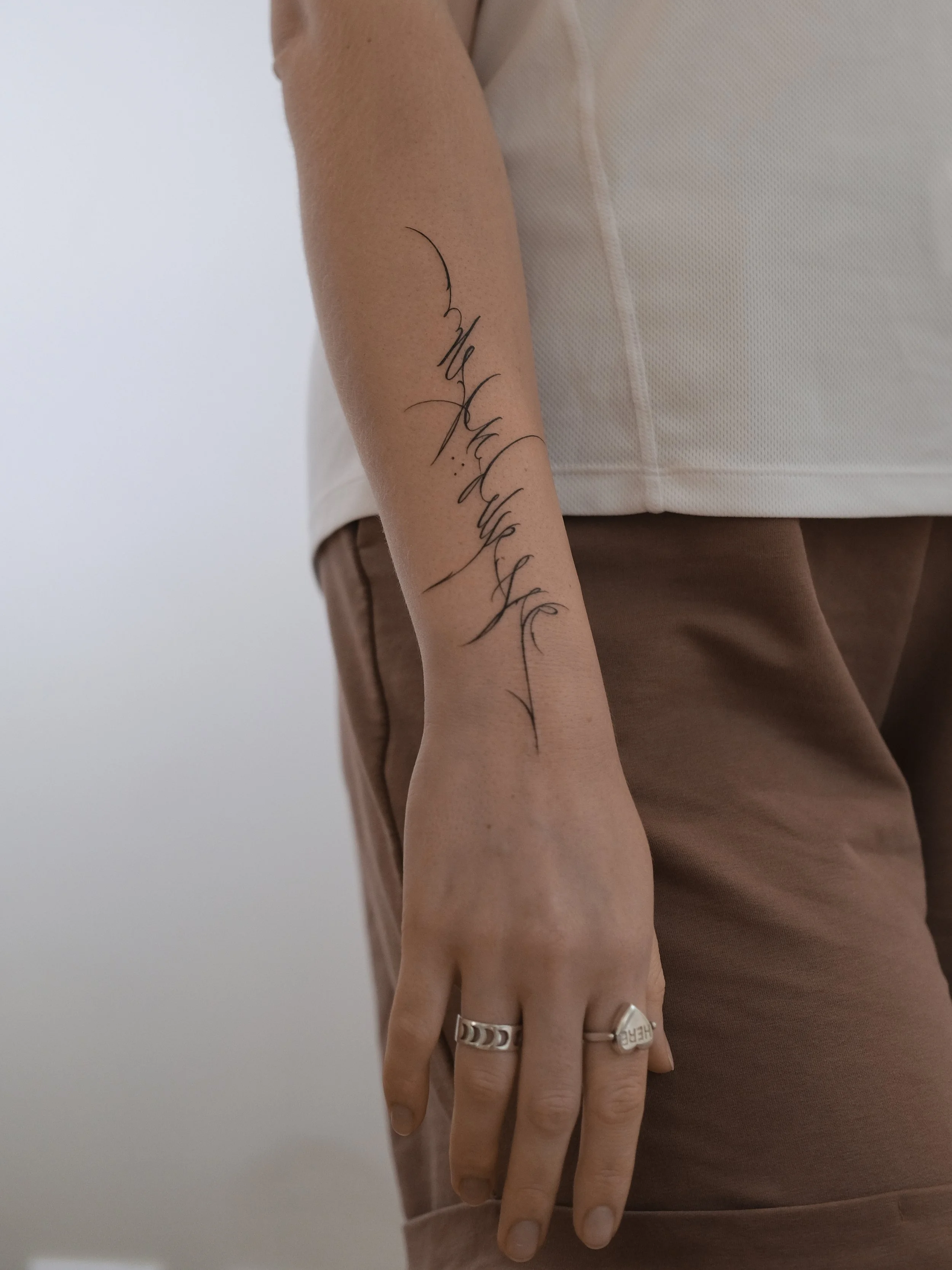 A person's arm with a tattoo and multiple rings, wearing a beige sleeveless top and brown pants, against a plain white background.