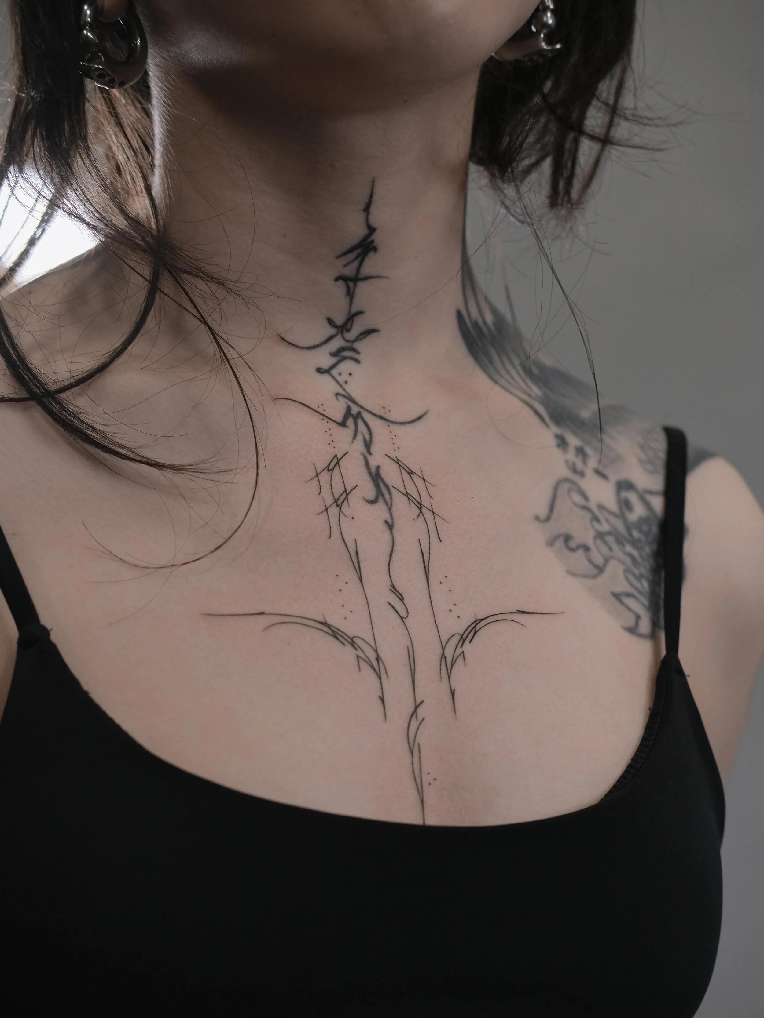 Close-up of a woman's neck with a minimalist black line tattoo of a girl falling, her arms outstretched and hair flowing, on the front of her neck, near her collarbone.
