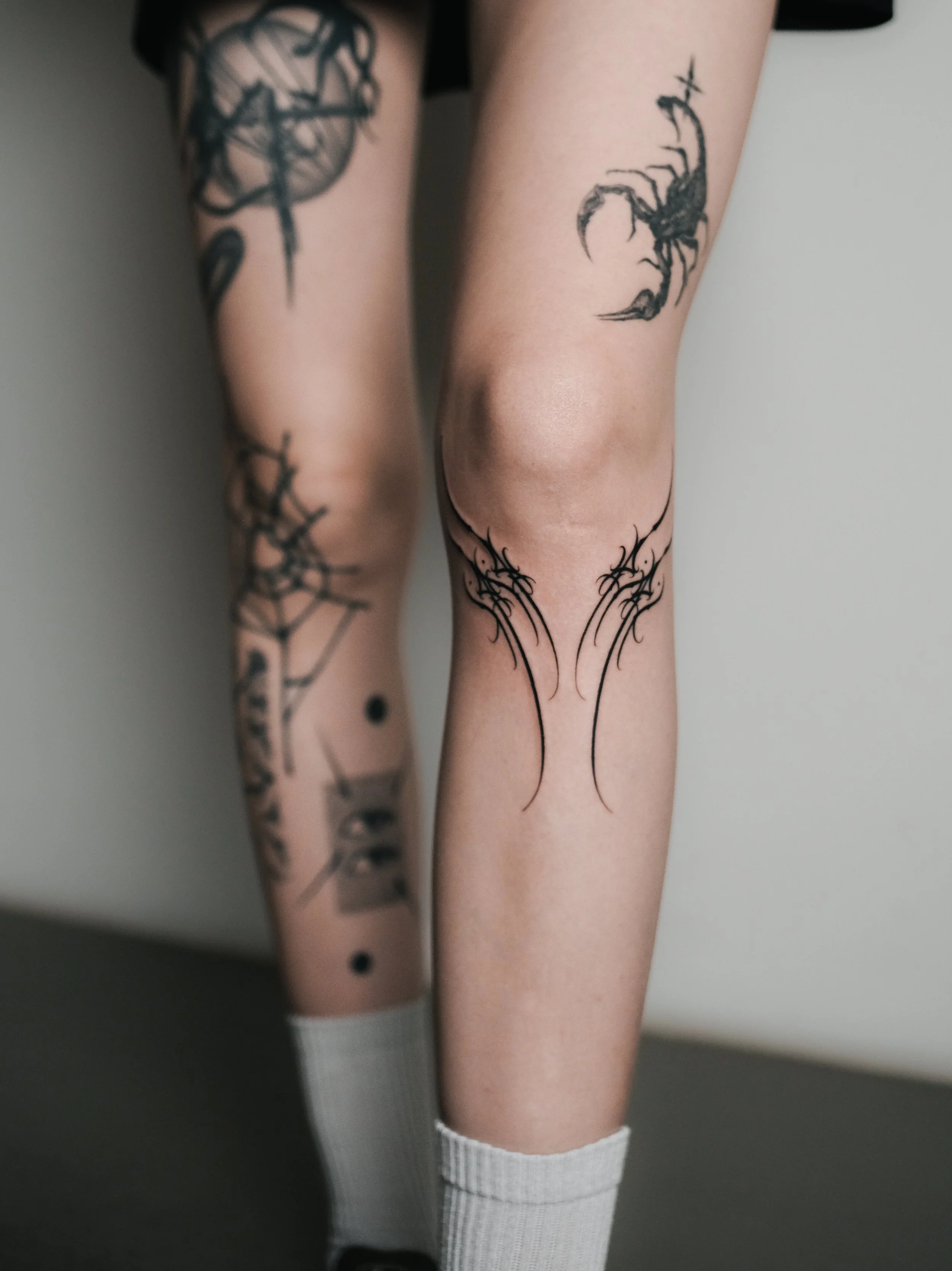 Close-up of two person's legs with tattoos. One leg has a scorpion tattoo and the other has a sword tattoo. Both are above the knee, and the person with the tattoos is wearing white knee-high socks.