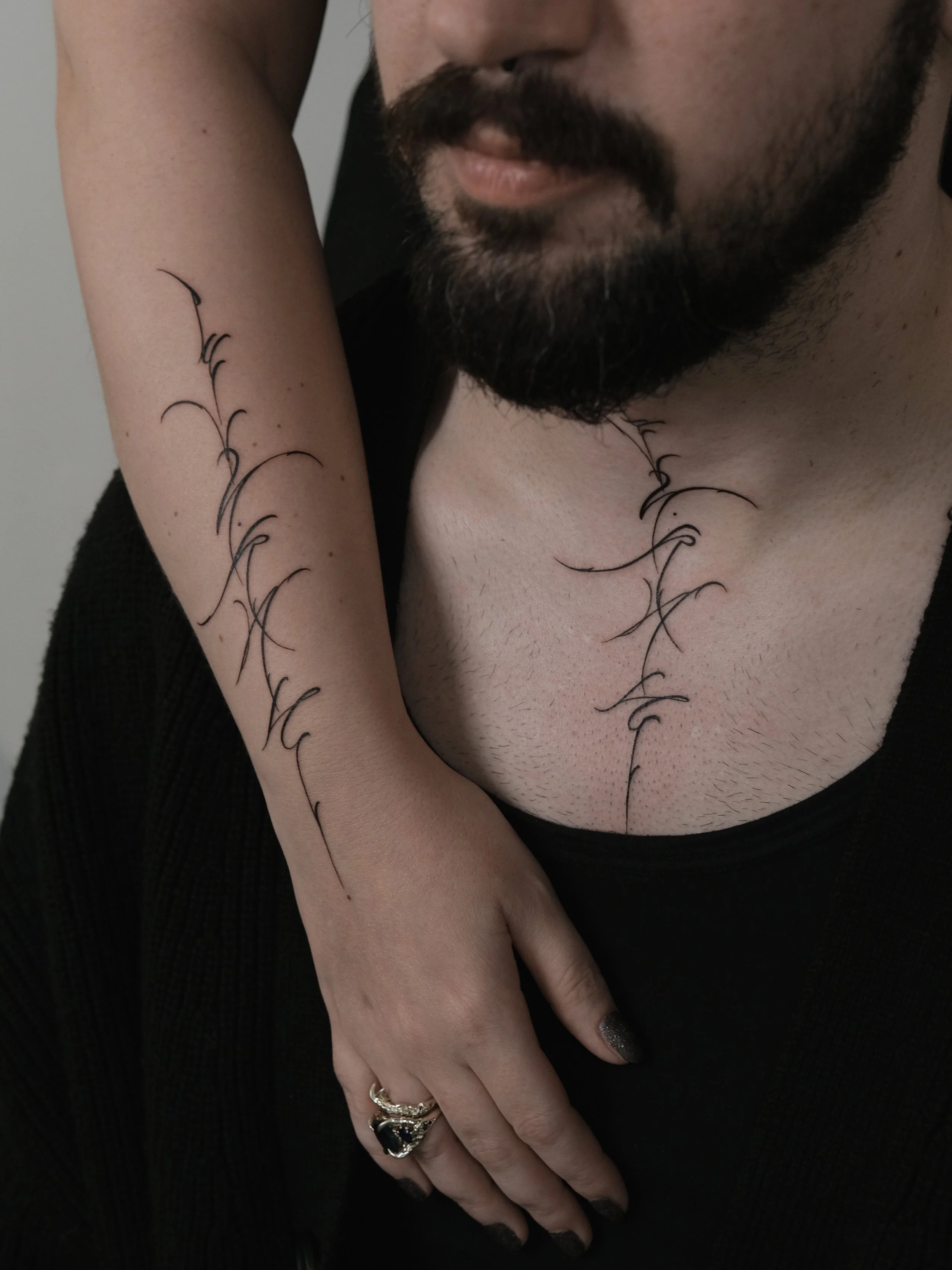 A person with tattoos on their forearm and chest, wearing rings and painted nails, with a beard and mustache, in a black sleeveless top.