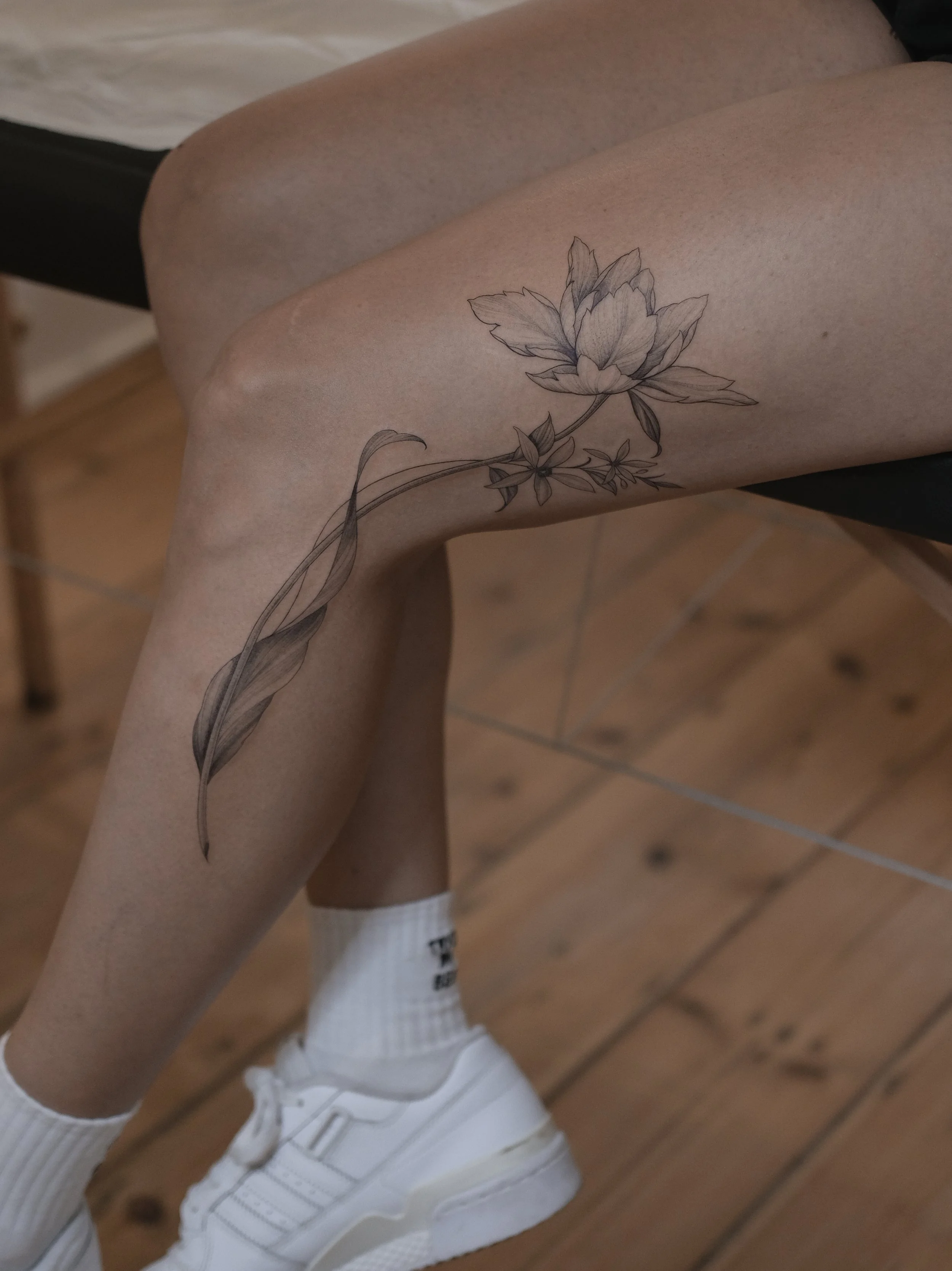 Close-up of a person's legs with a detailed black and white floral tattoo on one thigh, wearing white sneakers and socks, with a wooden floor background.
