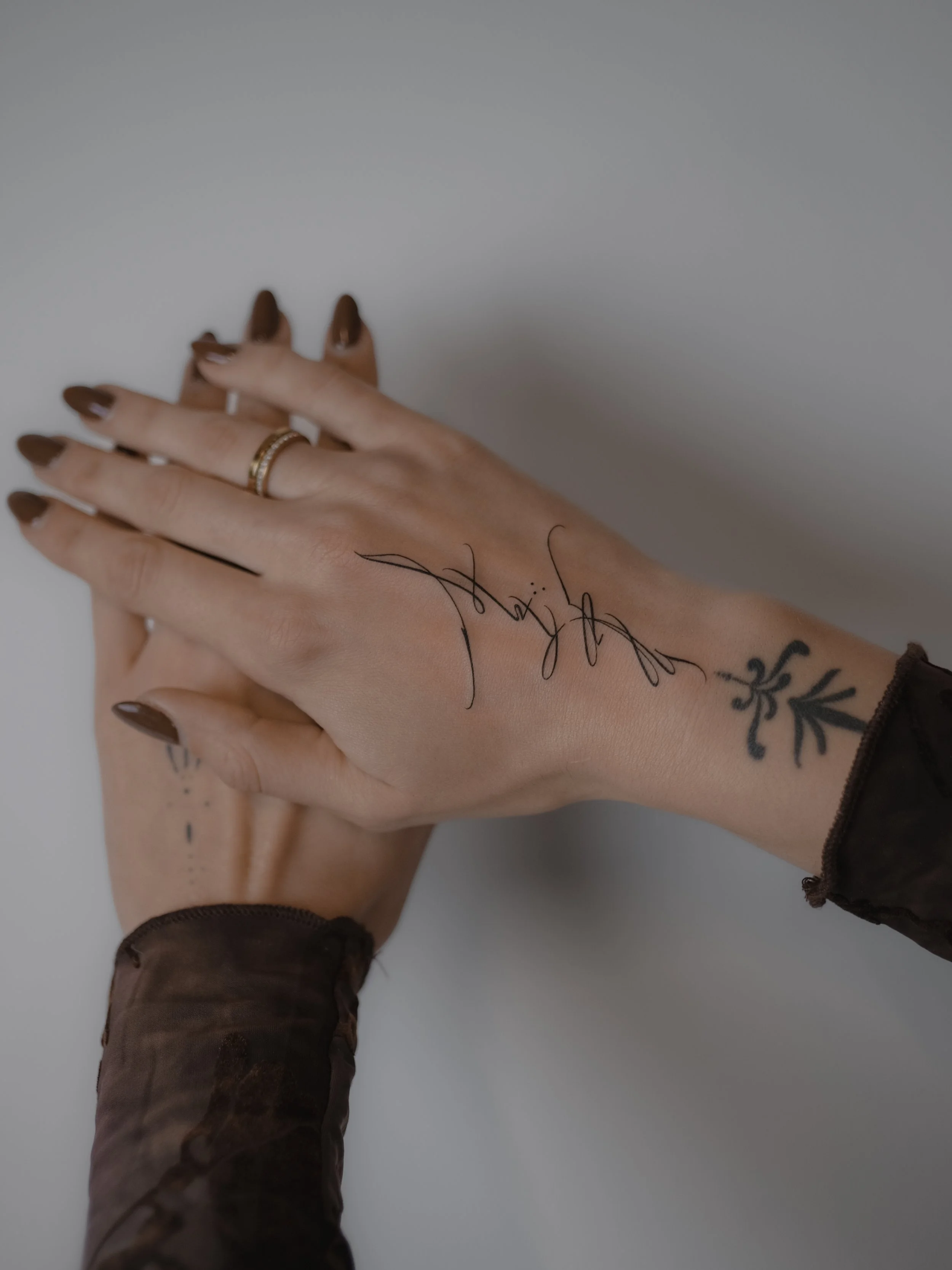 Close-up of two hands with tattoos and rings, holding each other against a plain light background.