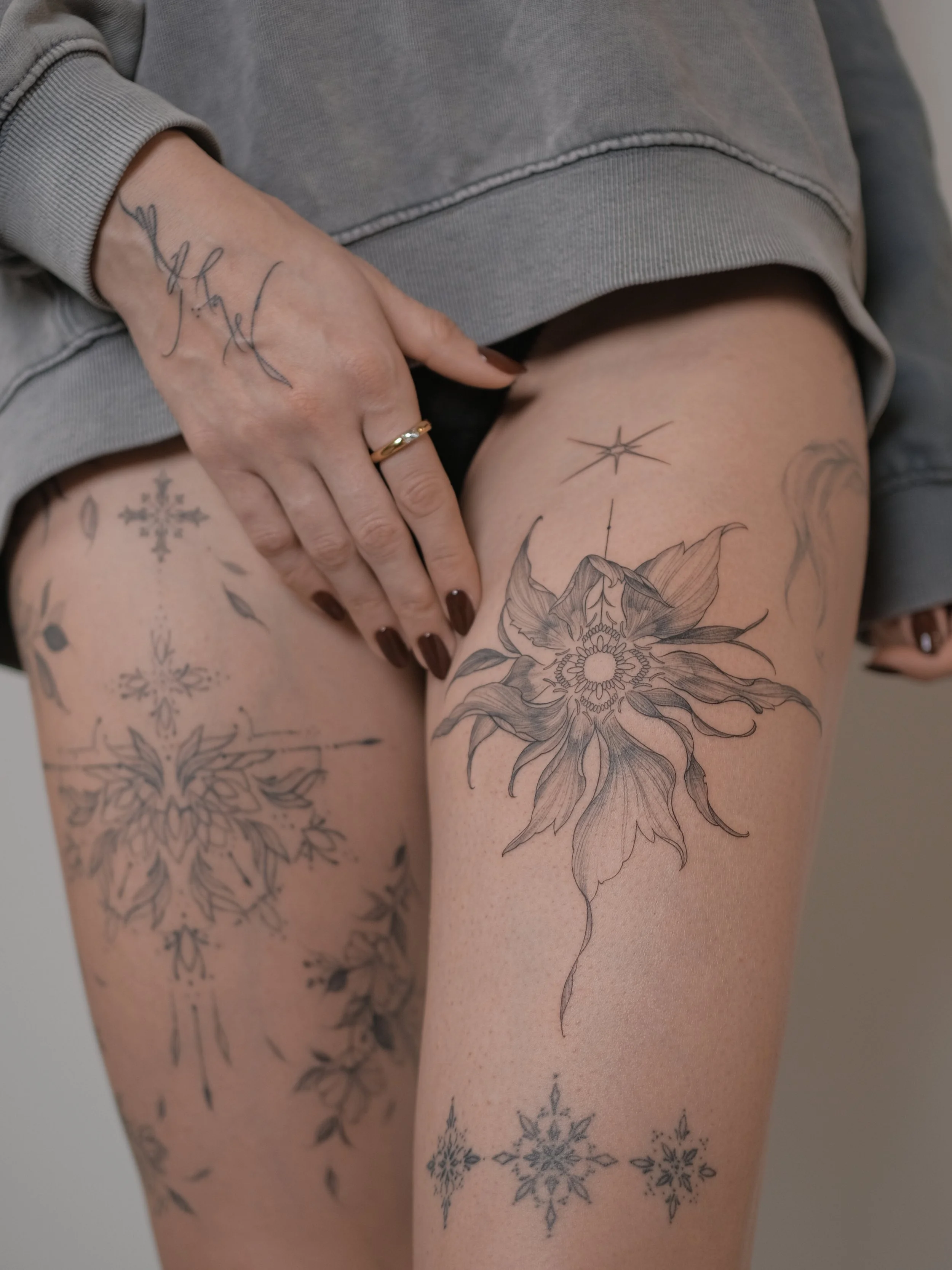 A person's thigh and hand showing various tattoos, including snowflakes, flowers, and a compass star, with the person wearing a gray sweatshirt and a gold ring.