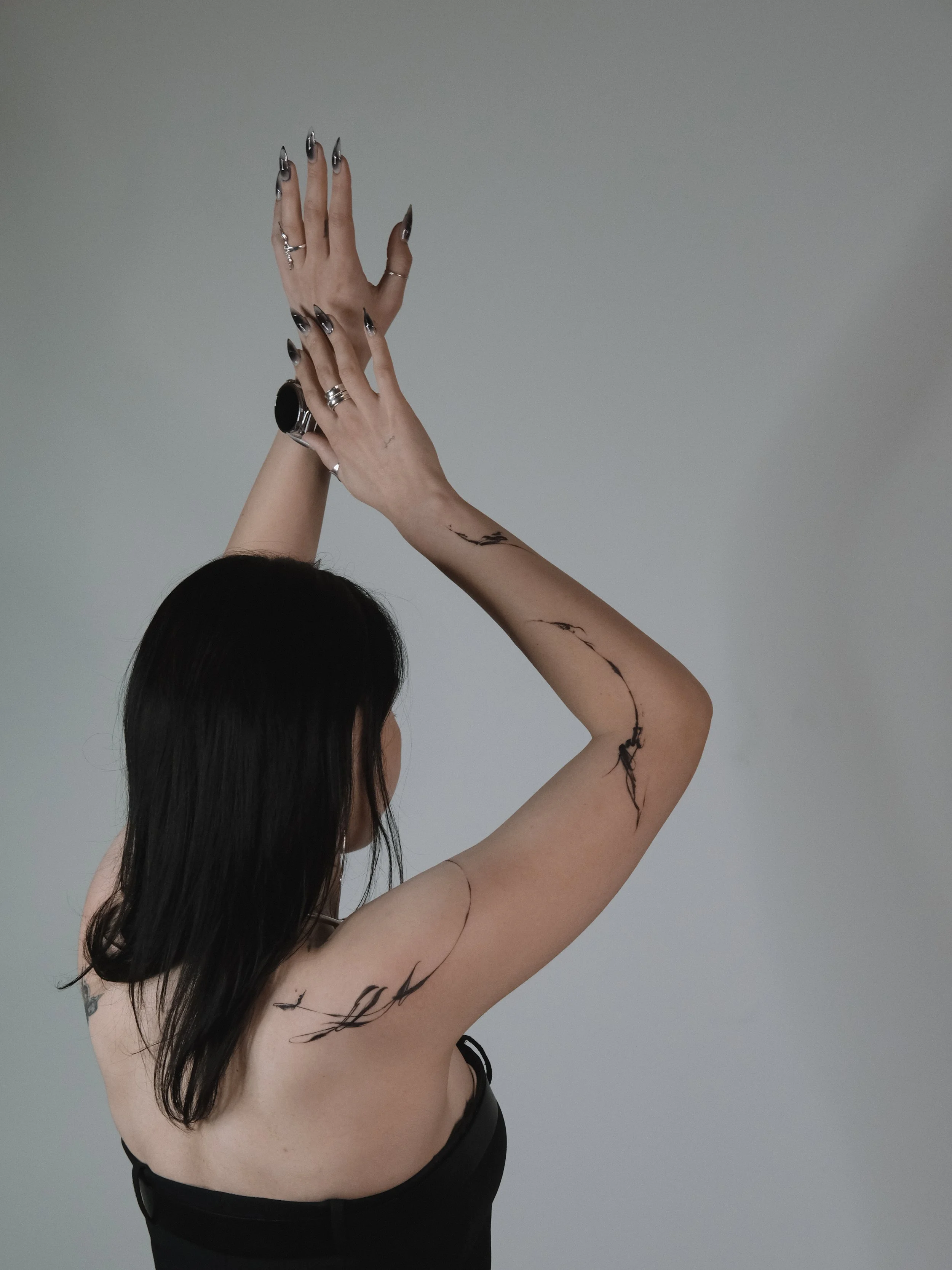 A woman with long dark hair, visible tattoos on her arm and shoulder, and multiple rings and rings on her fingers, holding her long fingers upward near her face against a plain gray background.