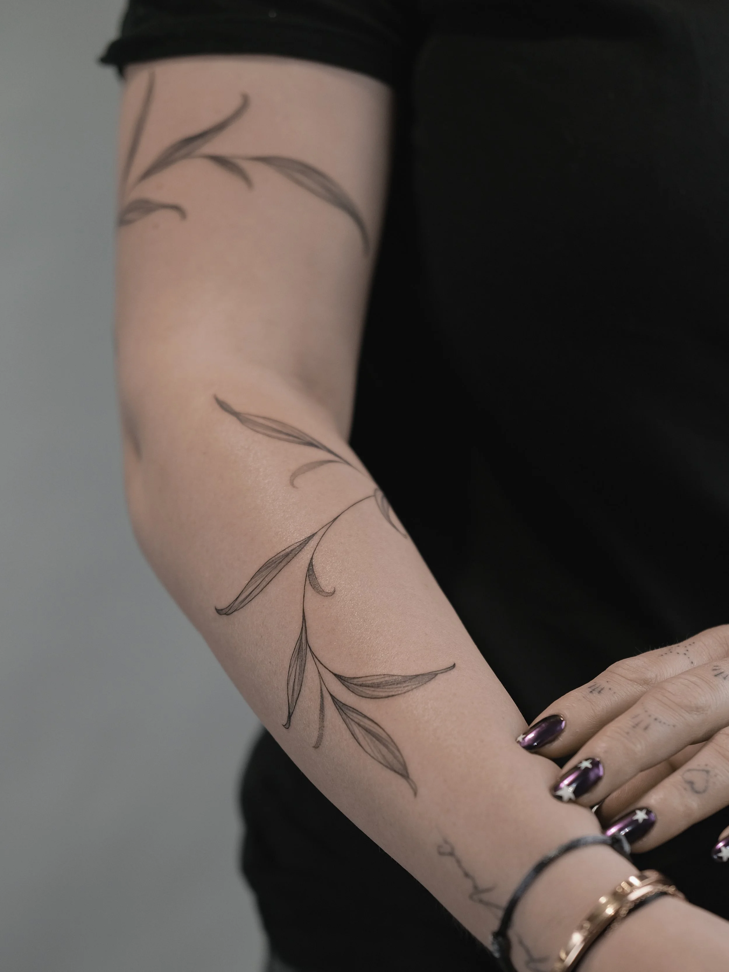 Close-up of a person's arm with a delicate black ink tattoo of a leafy vine, wearing dark nail polish with star designs and multiple bracelets.