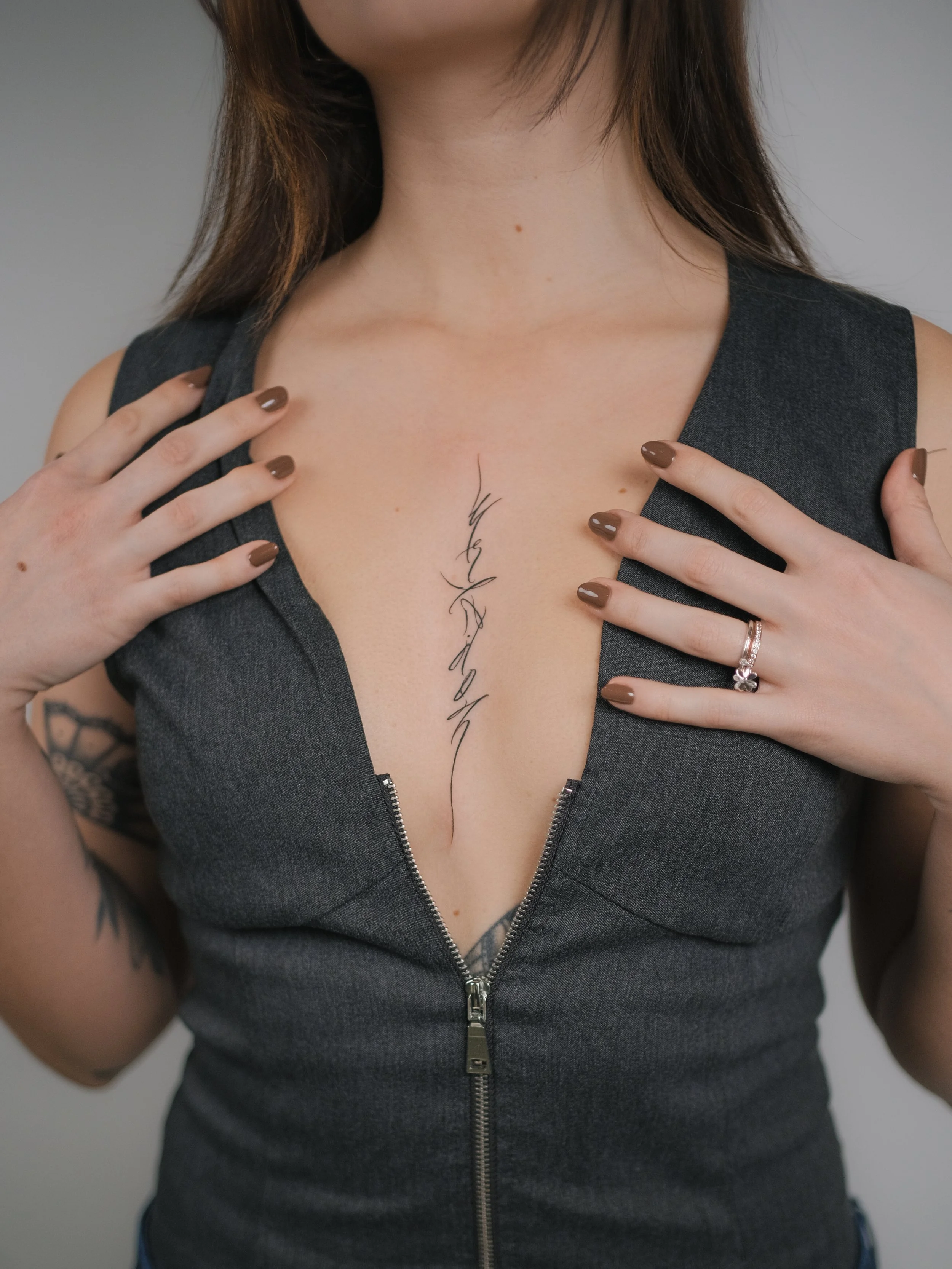 Close-up of a woman's neck and chest with a tattoo, wearing a dark gray sleeveless top with a zipper, touching her collarbone with both hands, fingernails painted brown, rings on fingers, and brown hair.