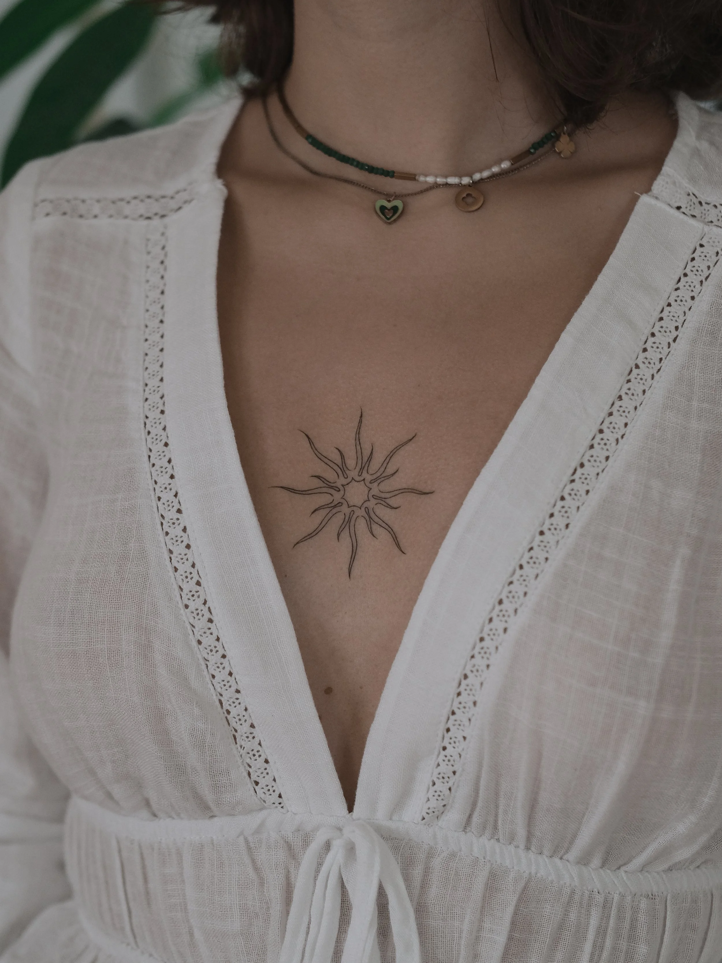 Close-up of a woman's chest and neck, showing layered necklaces and a tattoo of a sun or starburst design on her skin. She wears a white, embroidered, V-neck top with a drawstring at the waist.